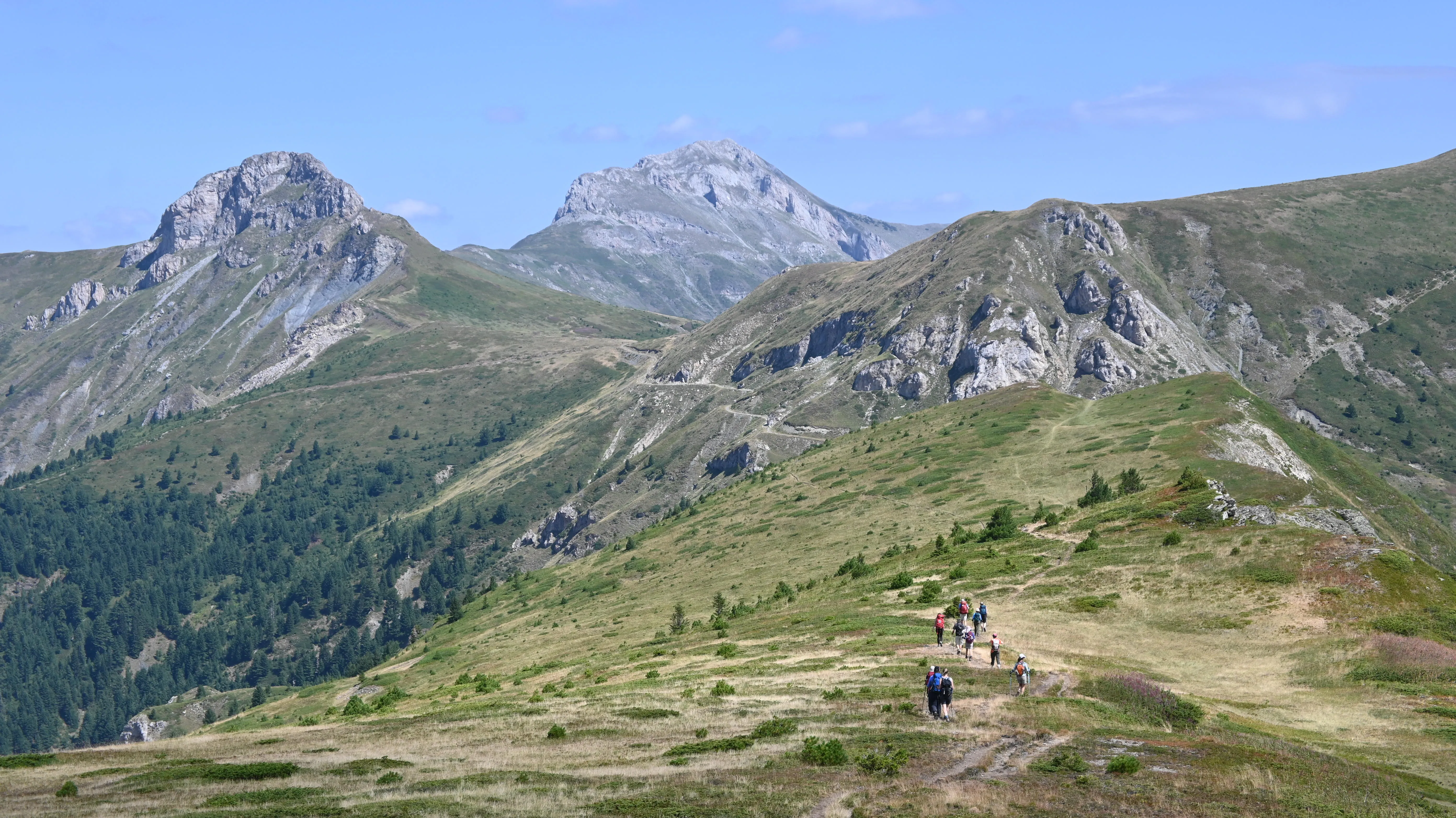 Hikers follow a winding trail across grassy ridges with panoramic mountain views along the Via Dinarica
