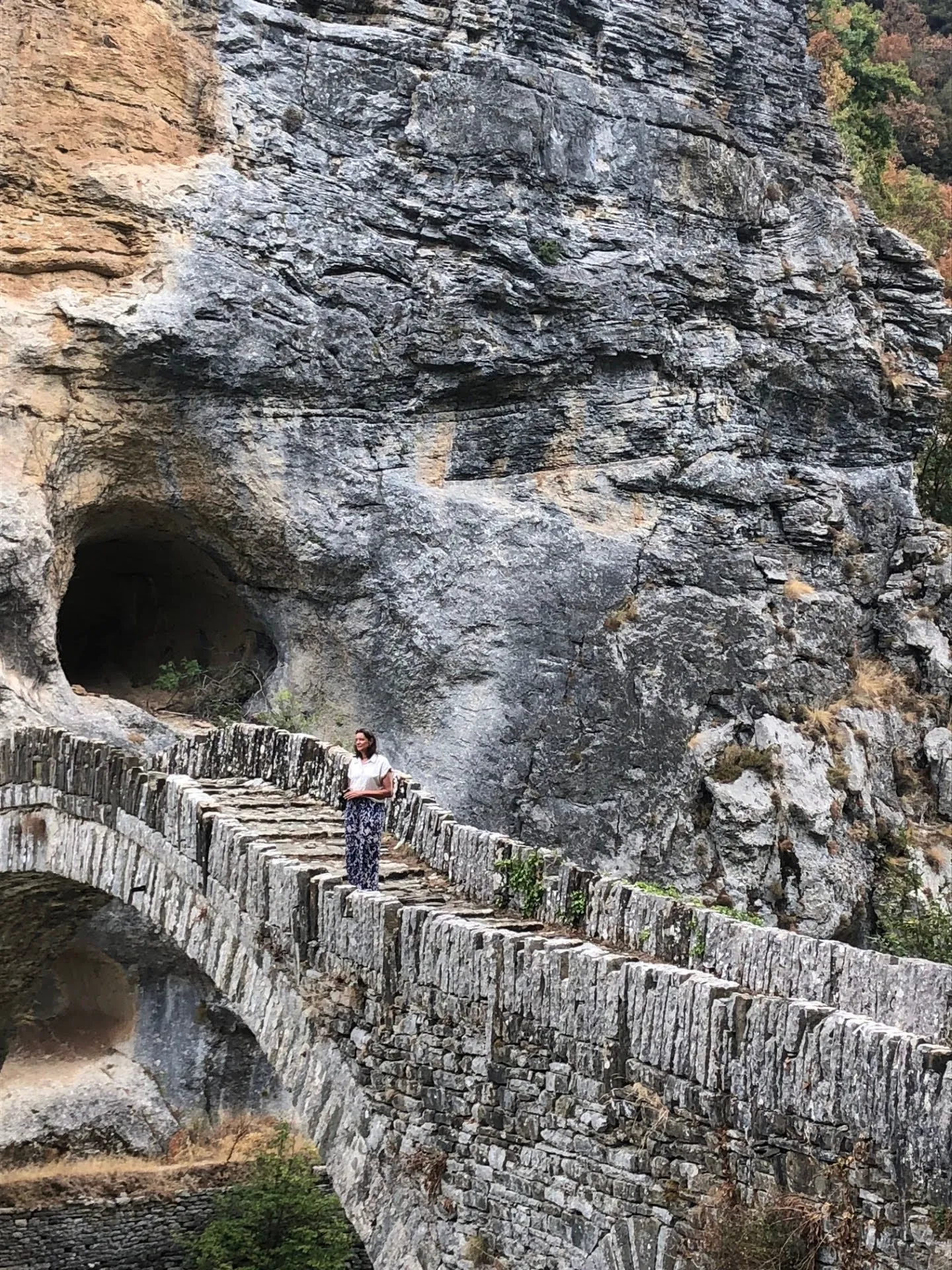 Traveler on Kokkorou Bridge, a historic stone arch bridge set against cliffs and caves in Zagori, Epirus.