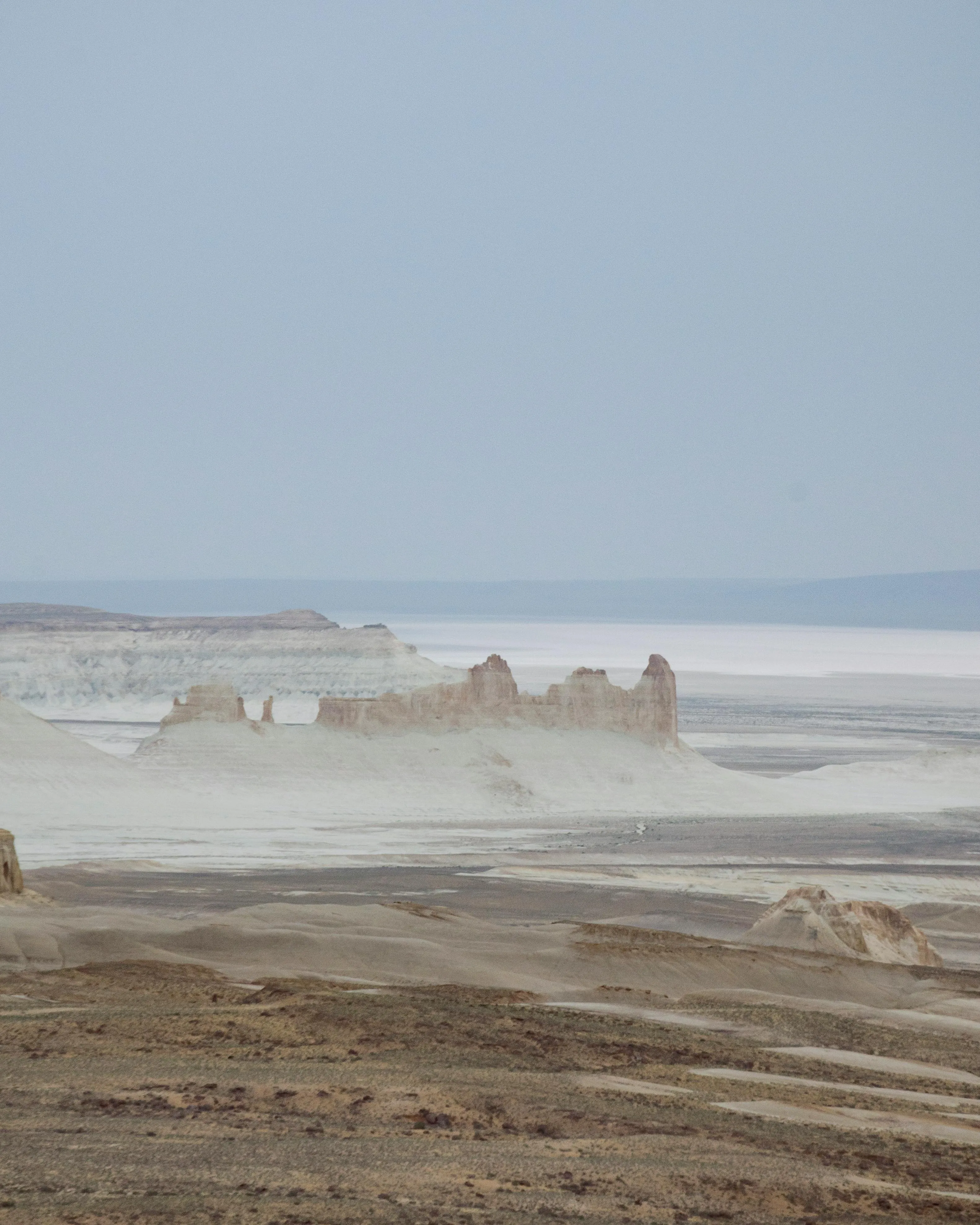 A rugged landscape in the Mangystau region of Kazakhstan, featuring towering rock formations and a vast desert-like terrain under a clear sky.