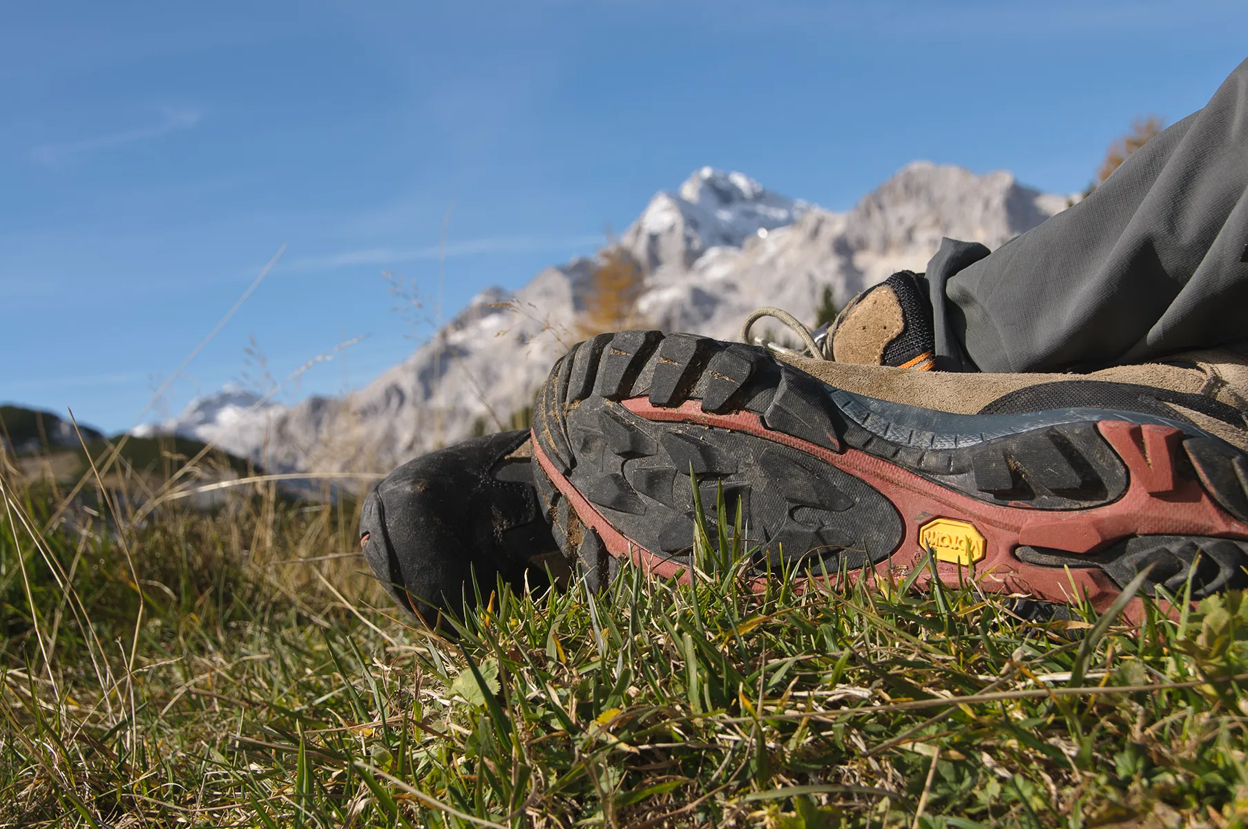 A pair of worn hiking boots resting on grass with a backdrop of the snow-capped peaks of the Julian Alps in Slovenia.