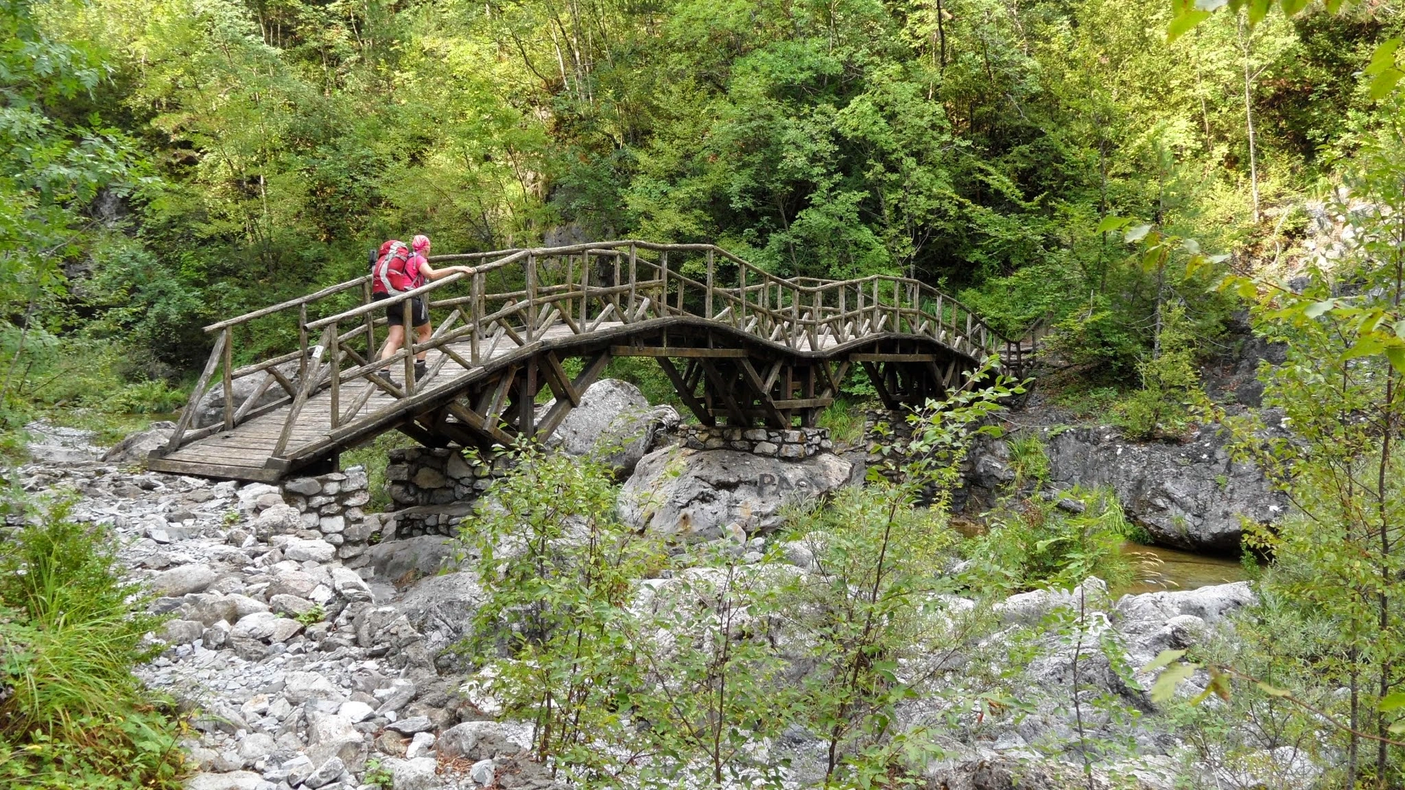 Hikers crossing a rustic wooden bridge over a rocky stream in Enippeus Gorge, Mount Olympus National Park, Greece.
