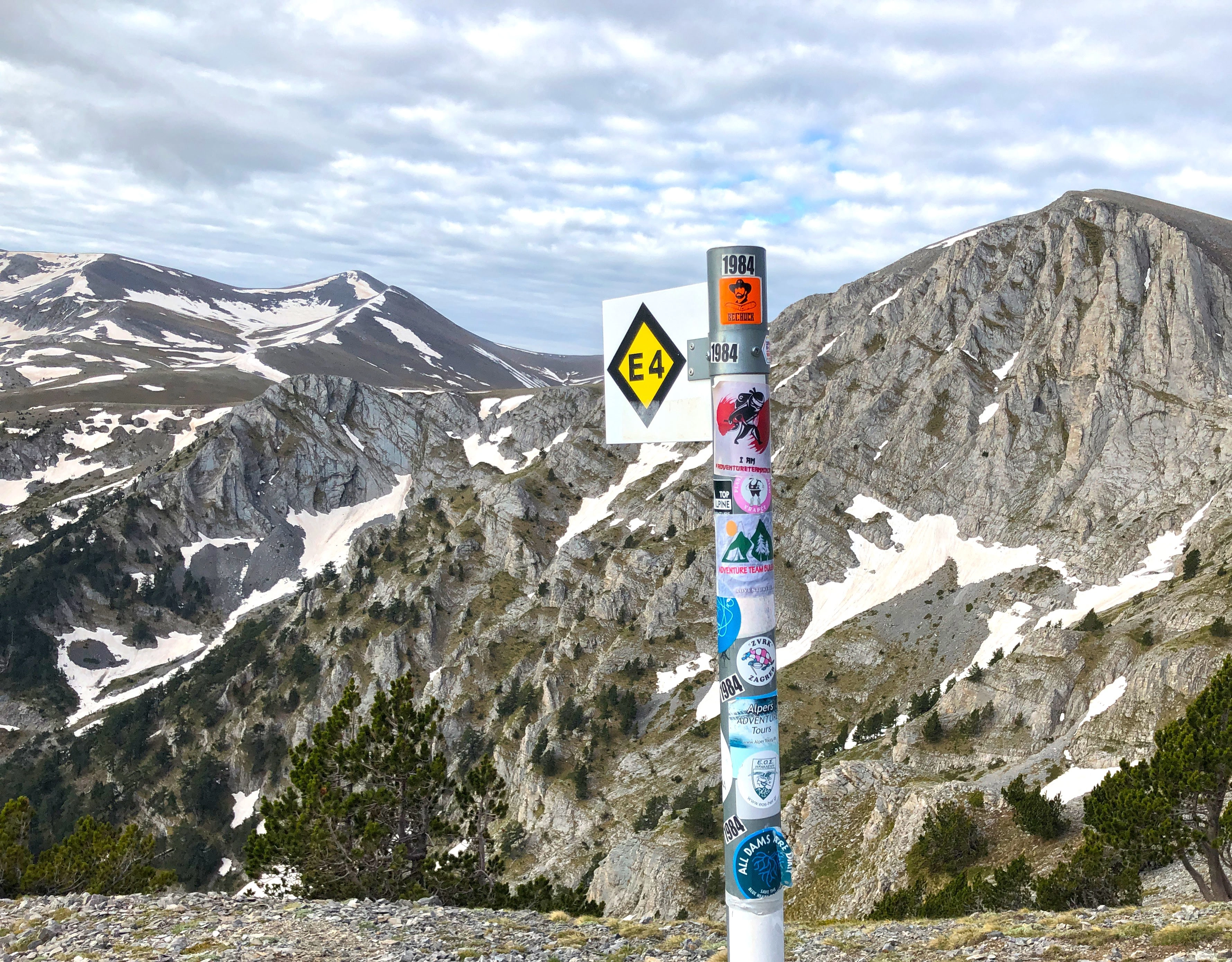 Signpost along the E4 European long-distance trail in Mount Olympus, Greece, with rugged mountain scenery and snow patches in the background.