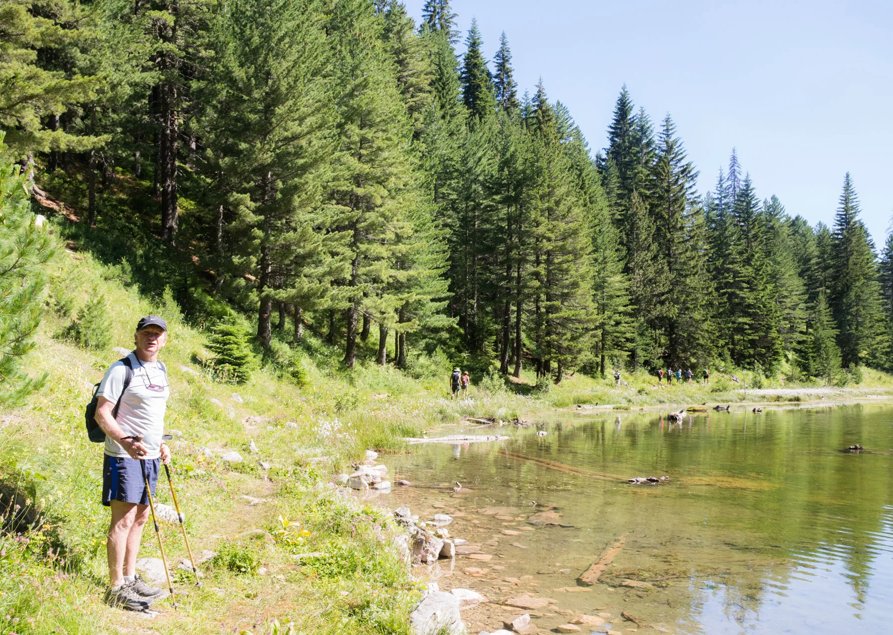 Hiker standing beside a forest lake on Kosovo’s Via Dinarica trail, with clear water and tall pines.