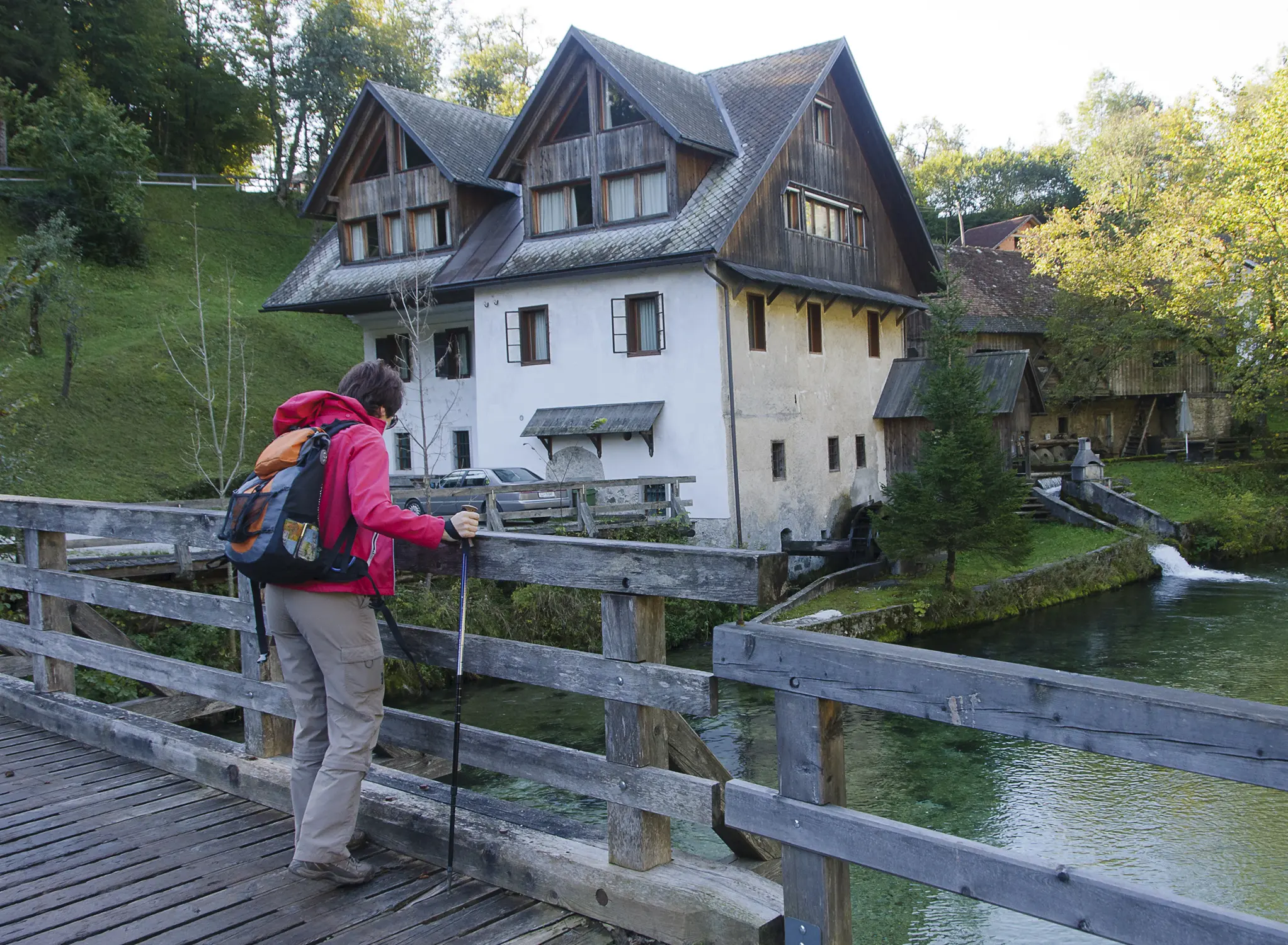 Hiker with trekking poles standing on wooden bridge, looking toward a rustic alpine house and watermill.