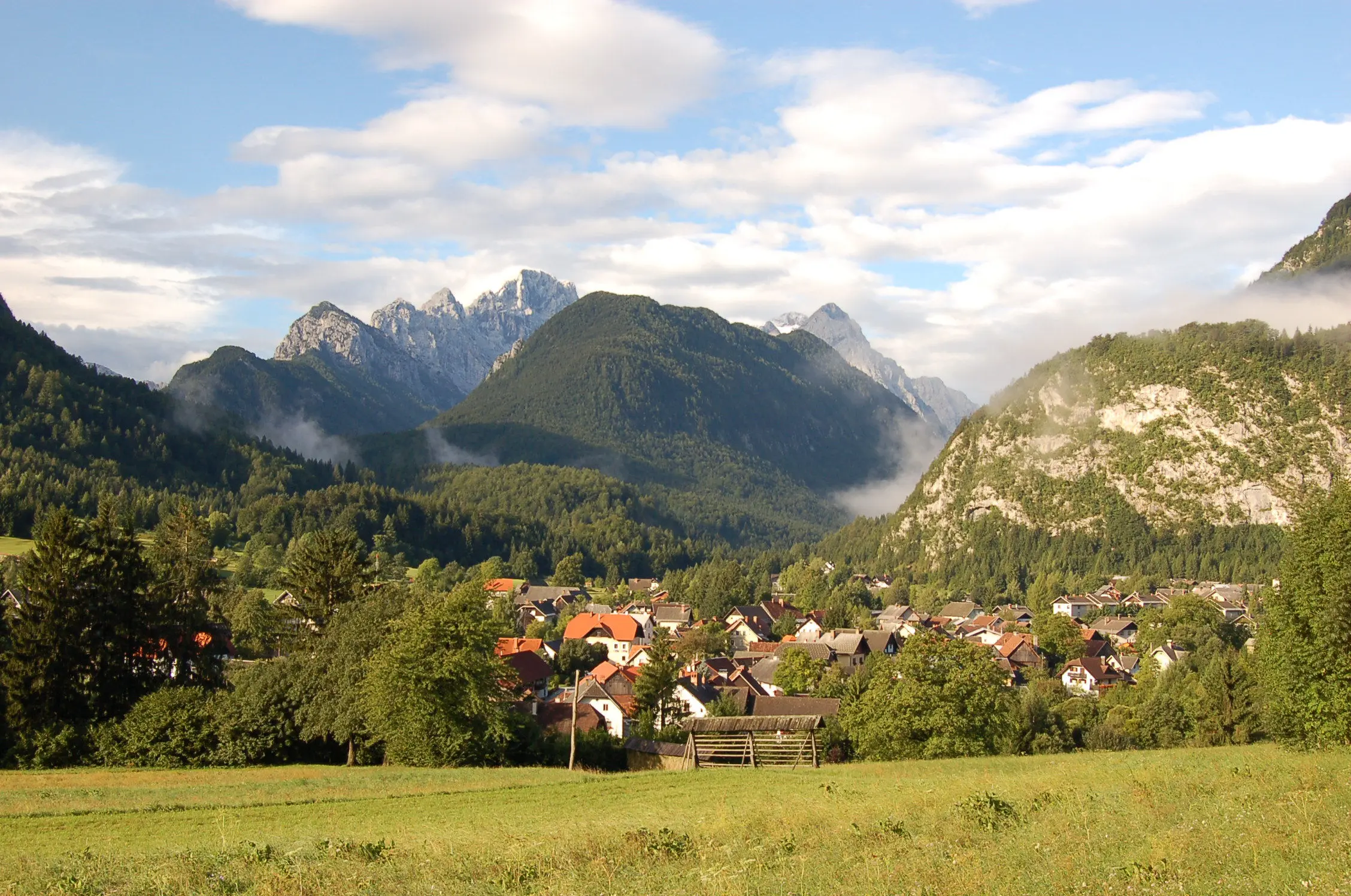 Alpine village with red-roofed houses nestled among green hills and dramatic mountain peaks in the background.