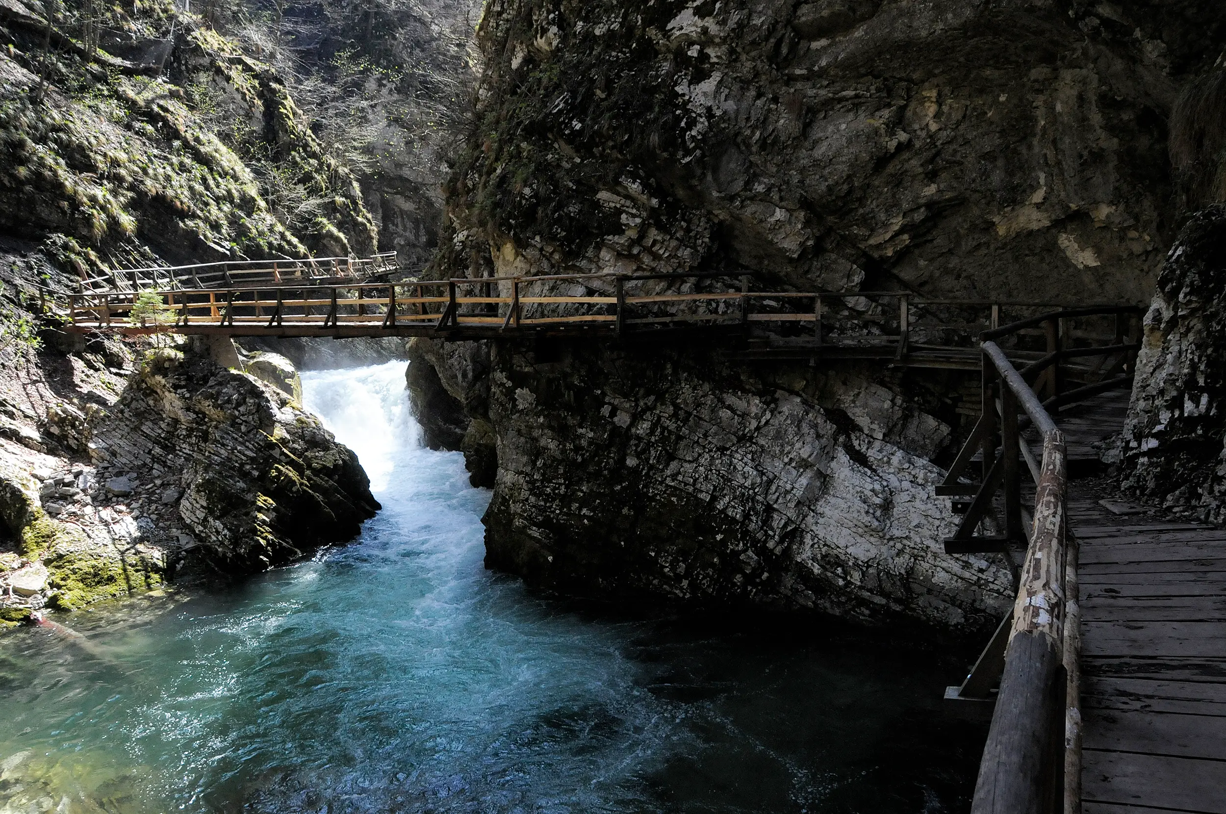 Wooden bridge crossing a turquoise river gorge, with steep rocky cliffs and moss-covered rocks.