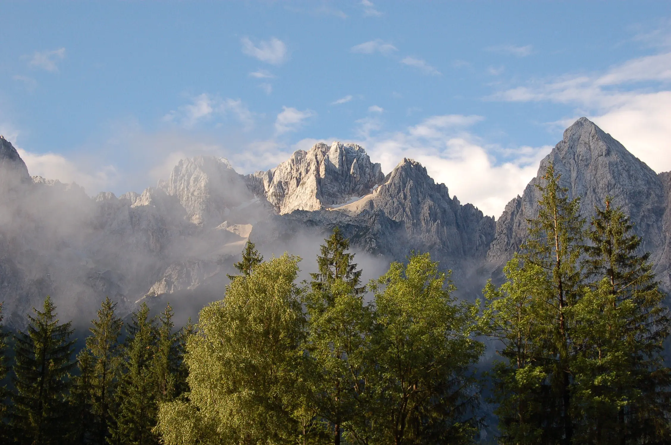 Jagged mountain peaks partially covered in mist, rising above lush green forests and clear blue skies.