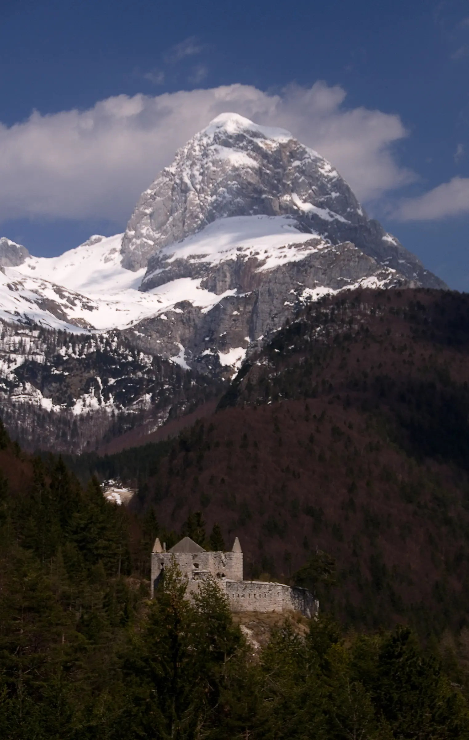Snow-capped mountain towering above forested hills, with a historic stone building nestled at the base.
