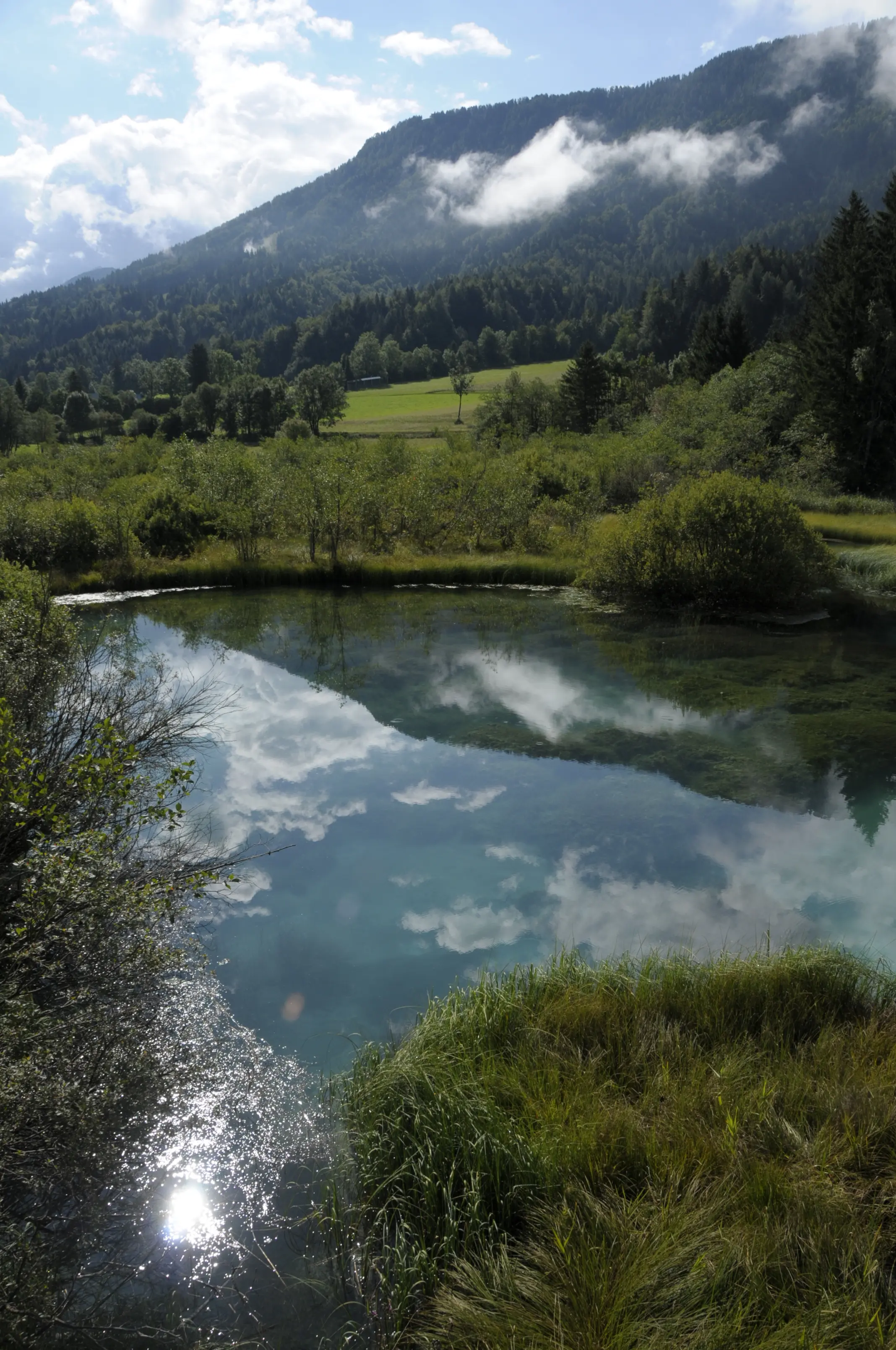 Crystal-clear pond reflecting sky and clouds, surrounded by green meadows and forested hills.