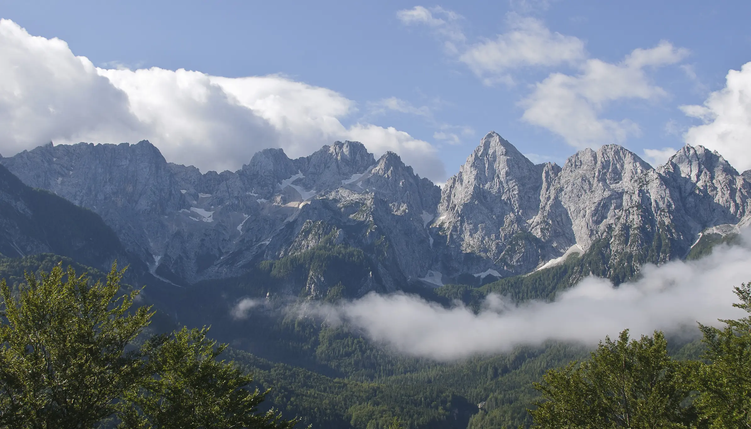 Low clouds drift across sharp peaks of the Julian Alps, framed by green forest under soft morning light.
