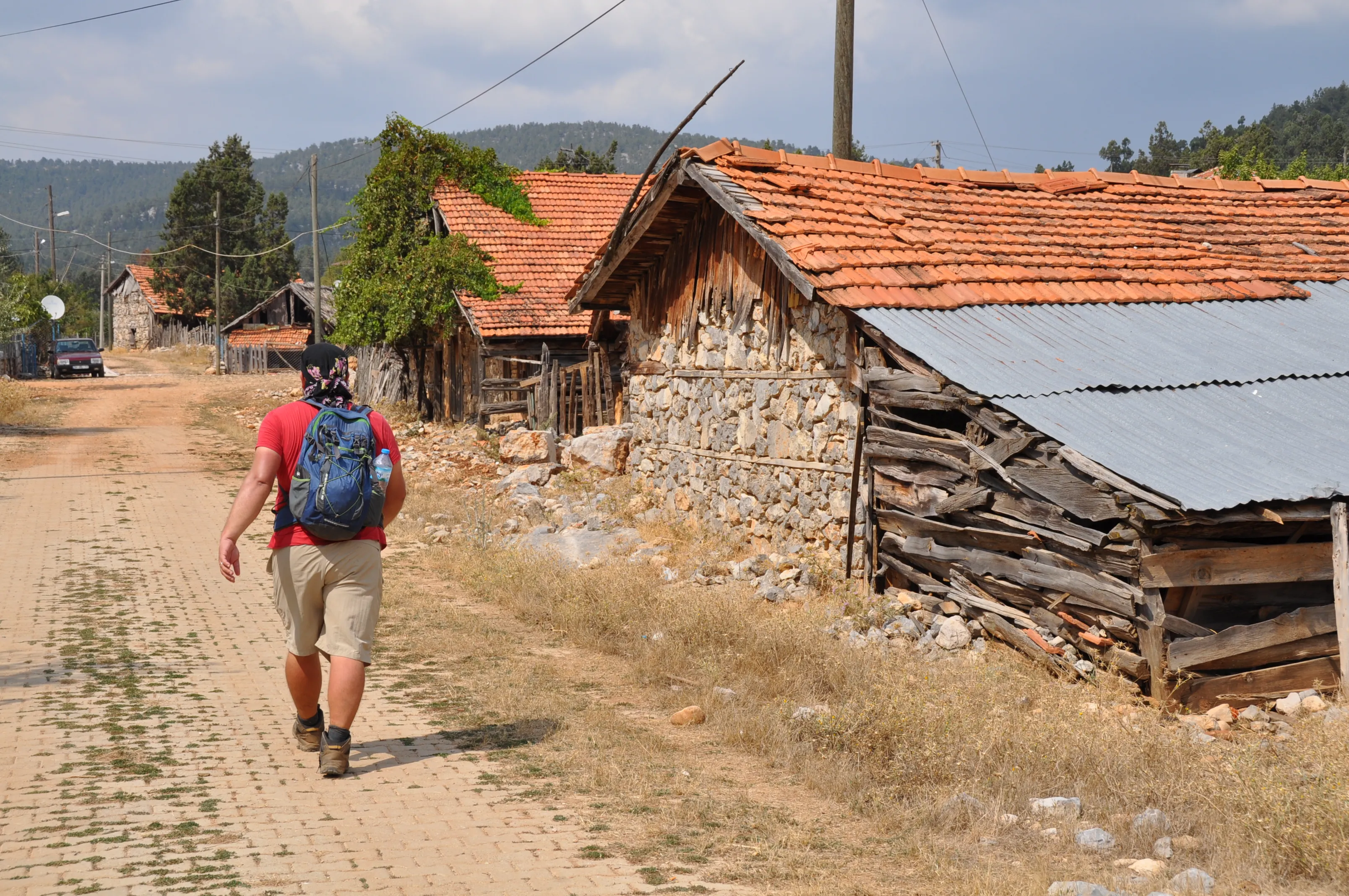 A hiker with a blue backpack walks past old stone houses with red-tiled roofs in a rustic village on the St. Paul Trail.