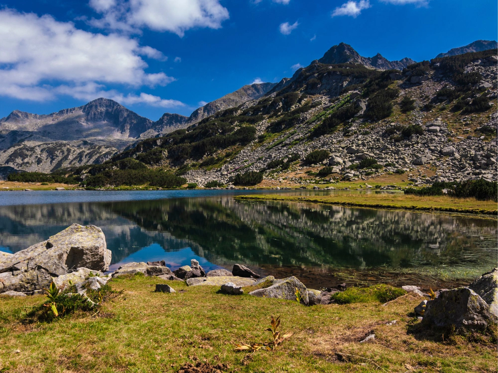 Crystal-clear lake in Pirin Mountain, Bulgaria, reflecting the surrounding rocky peaks and blue sky.