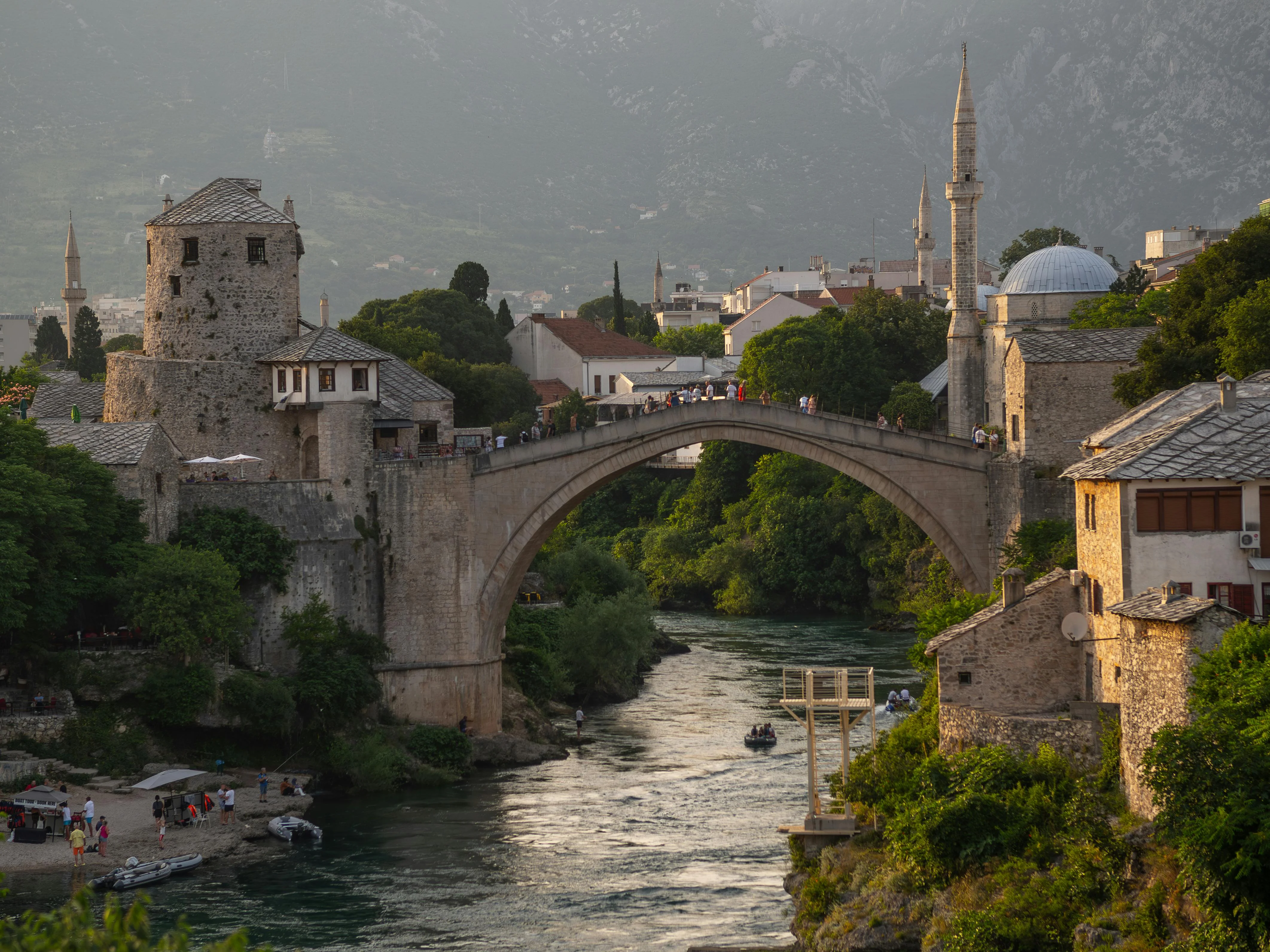 The historic Stari Most bridge in Mostar, Bosnia, at sunset, with people walking across and boats in the river below.