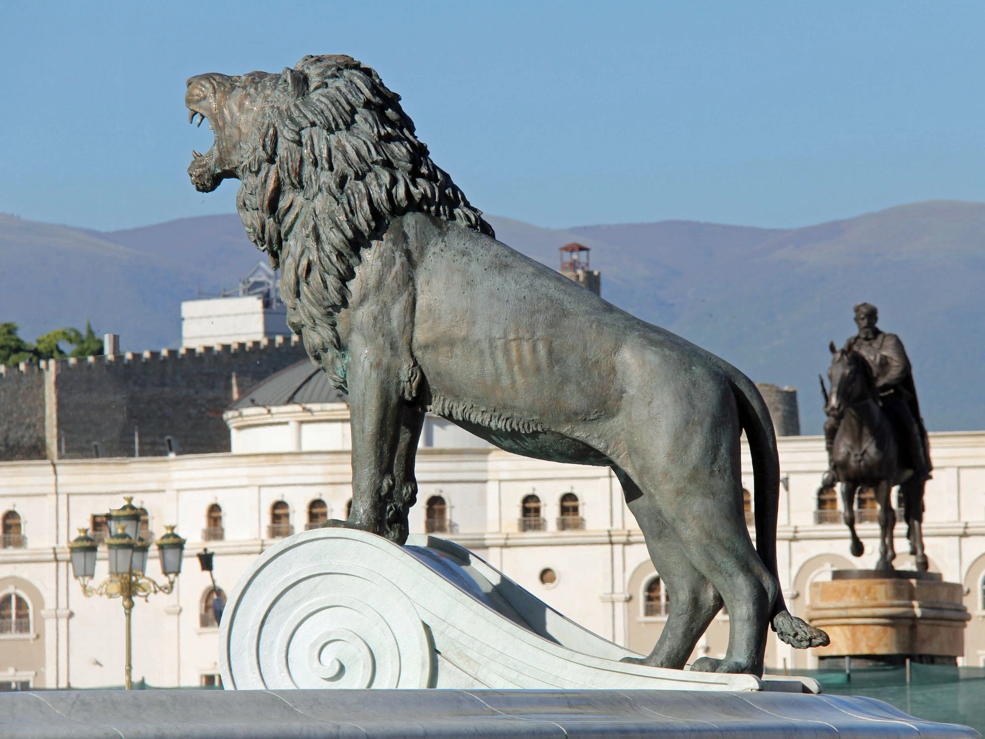 Skopje Lion – bronze lion statue roaring with mountain backdrop and equestrian statue behind.