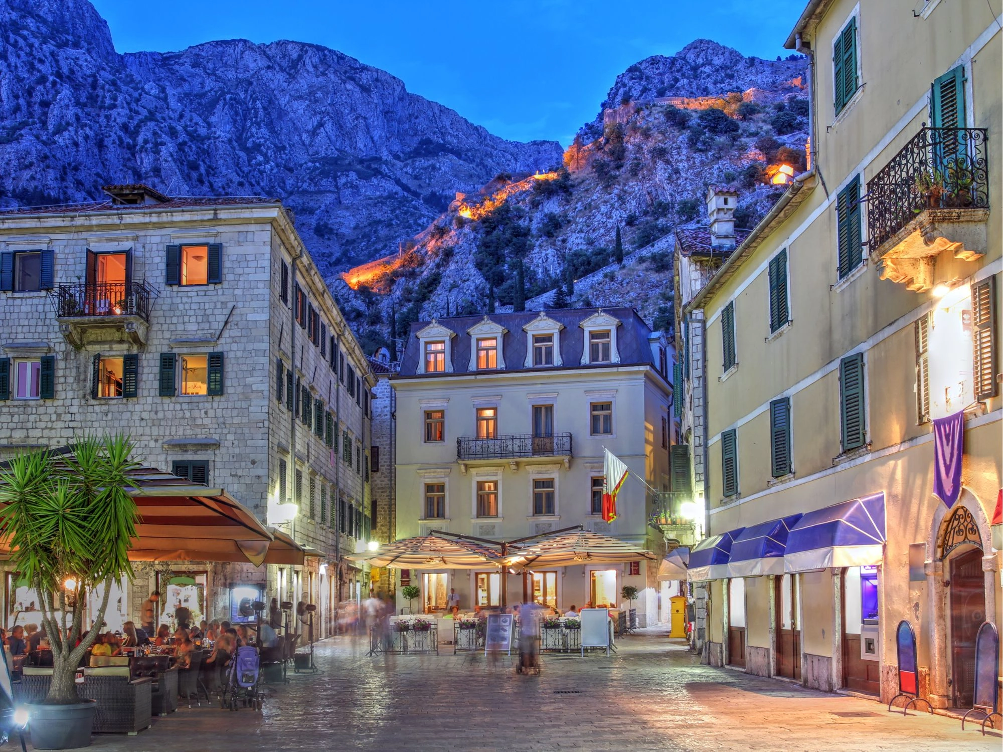 Montenegro – Kotor old town square at twilight with glowing windows and fortress on the hill.