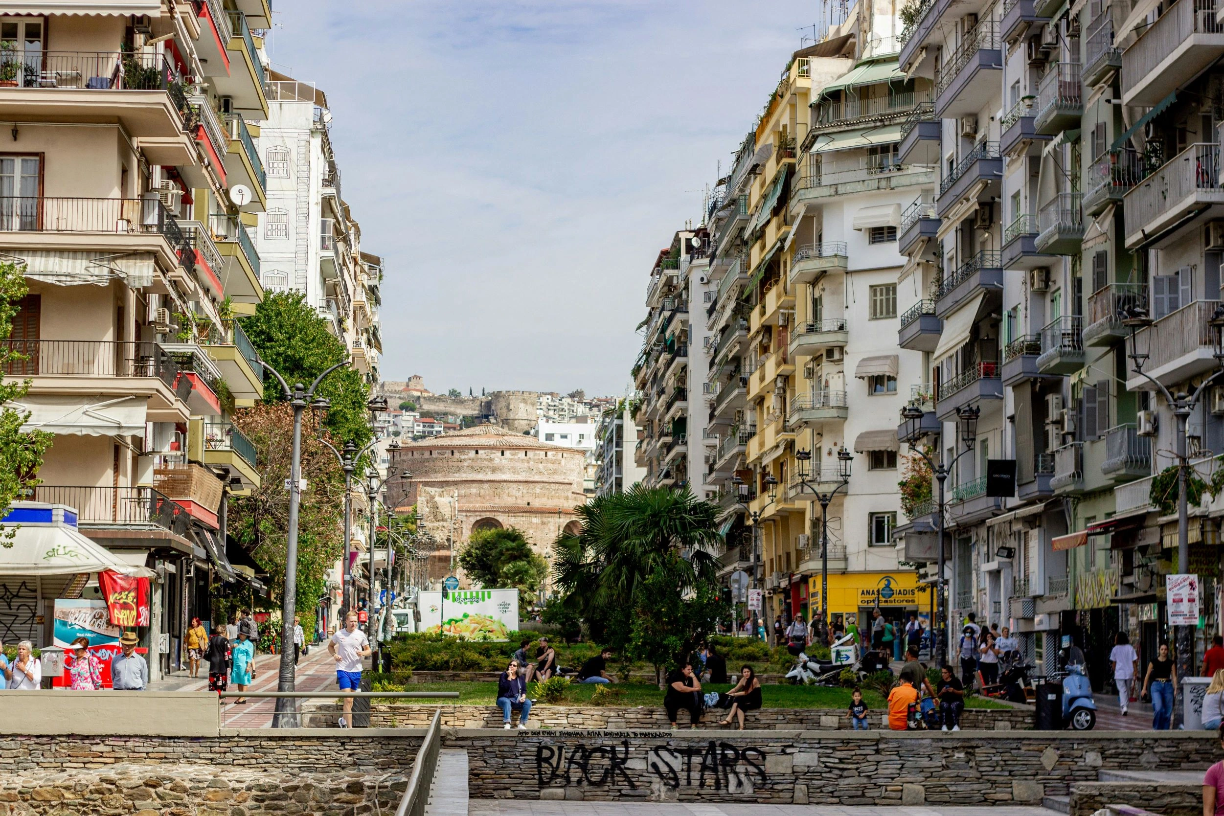Busy Thessaloniki street with Rotunda monument in distance and apartment buildings on both sides.Crowds walking in vibrant Thessaloniki with Rotunda monument visible in the distance.