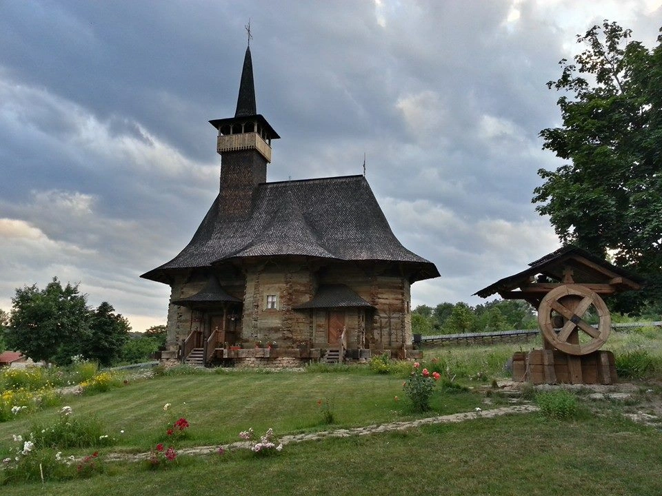 Old wooden church with steep shingle roof in Moldova, surrounded by wildflowers and a well.