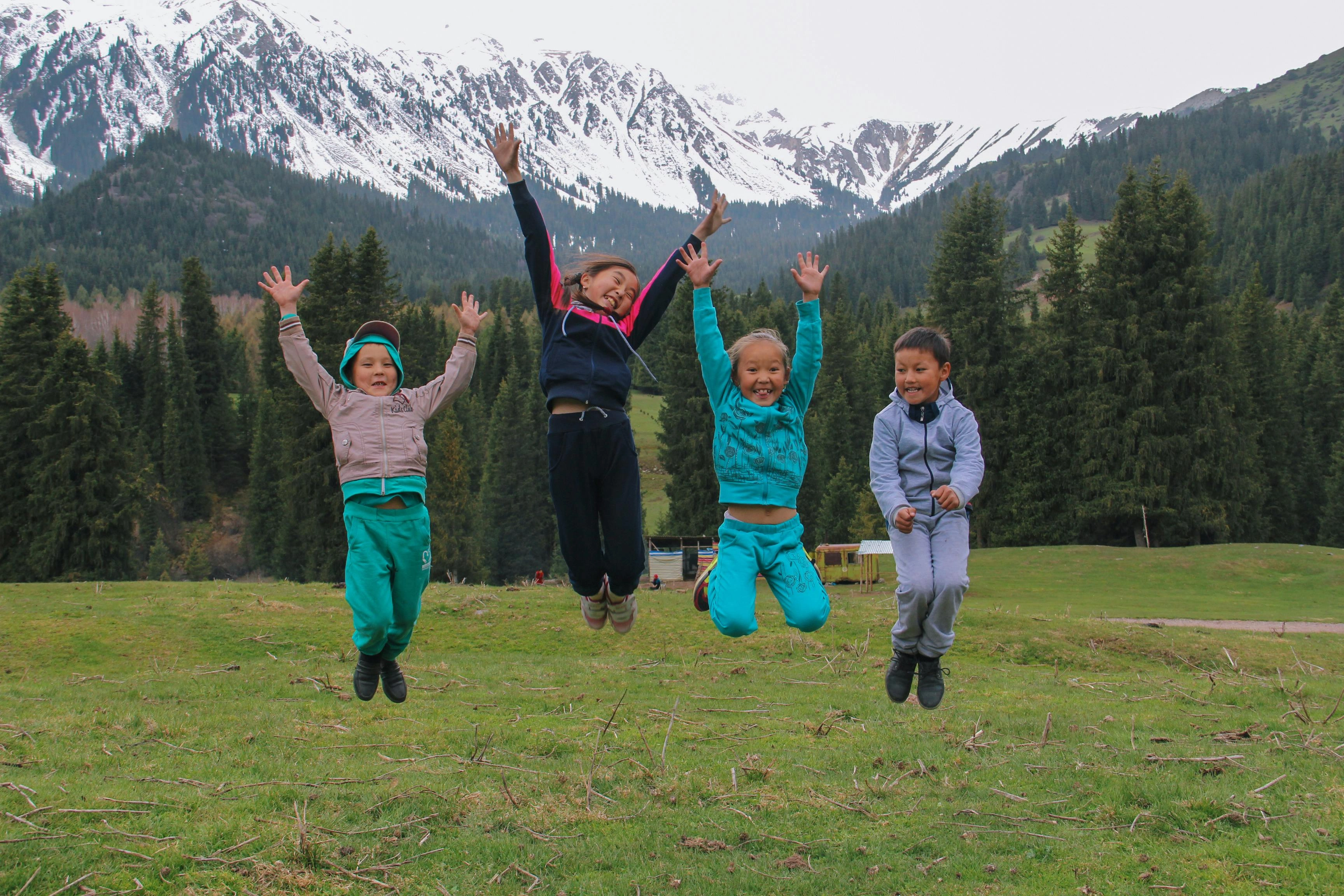 Group of children jumping joyfully in Kyrgyz meadow with snowy peaks and pine trees.