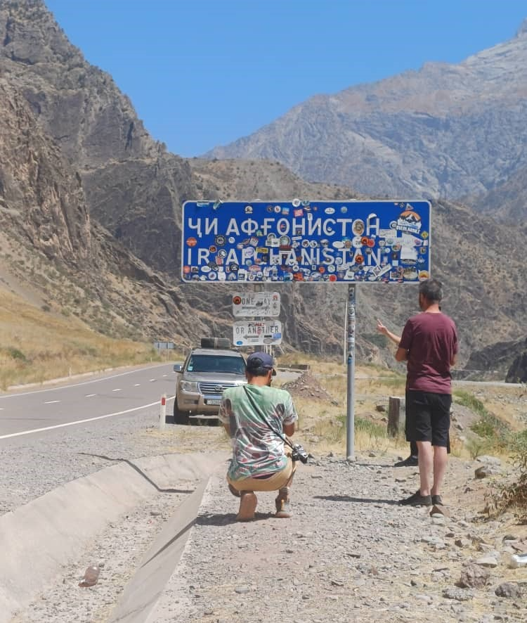 Tourists at the Tajik-Afghan border sign on the Pamir Highway, surrounded by rugged mountains.
