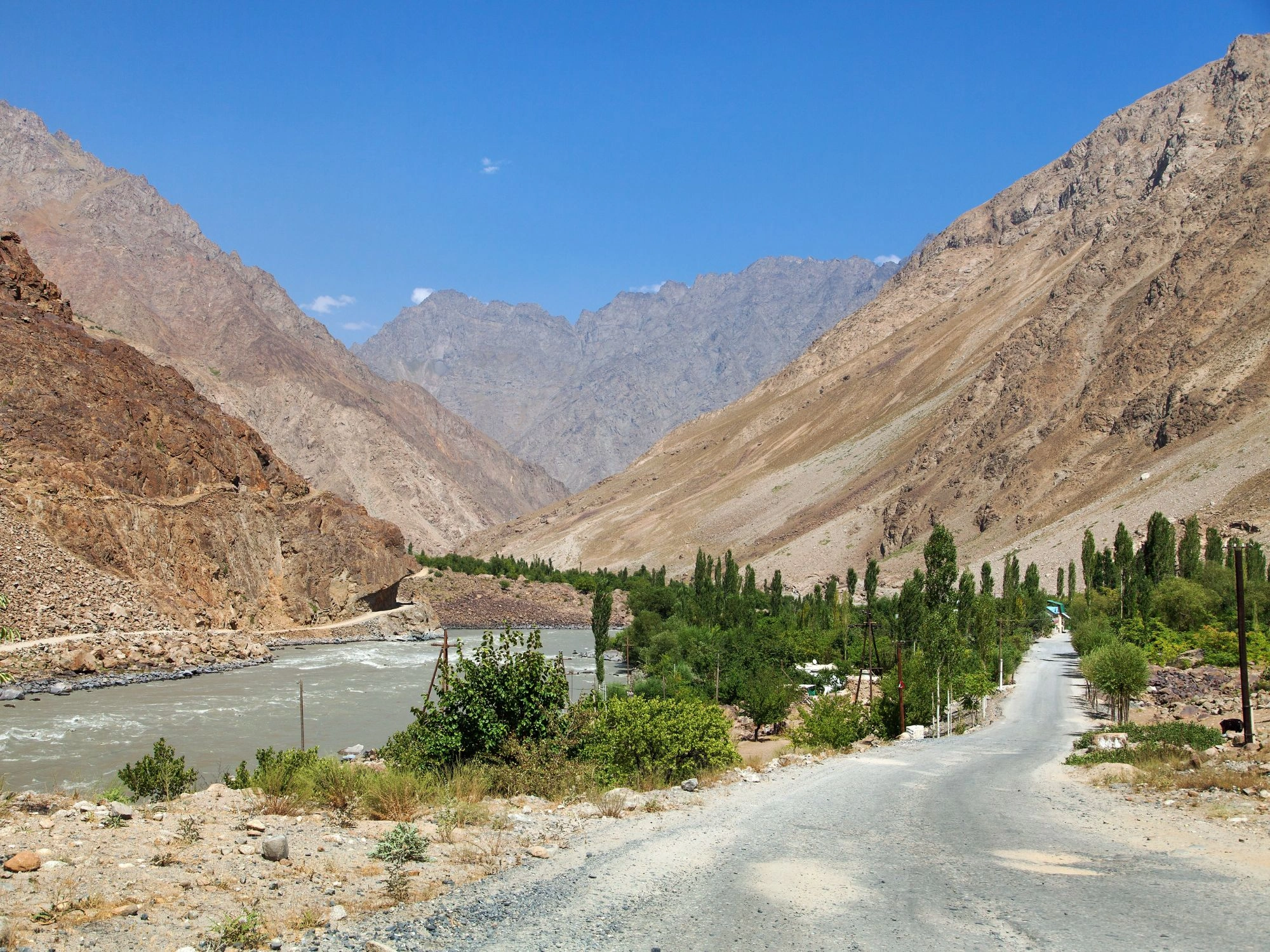 Gravel road runs beside river through green valleys and steep mountains on the Pamir Highway.