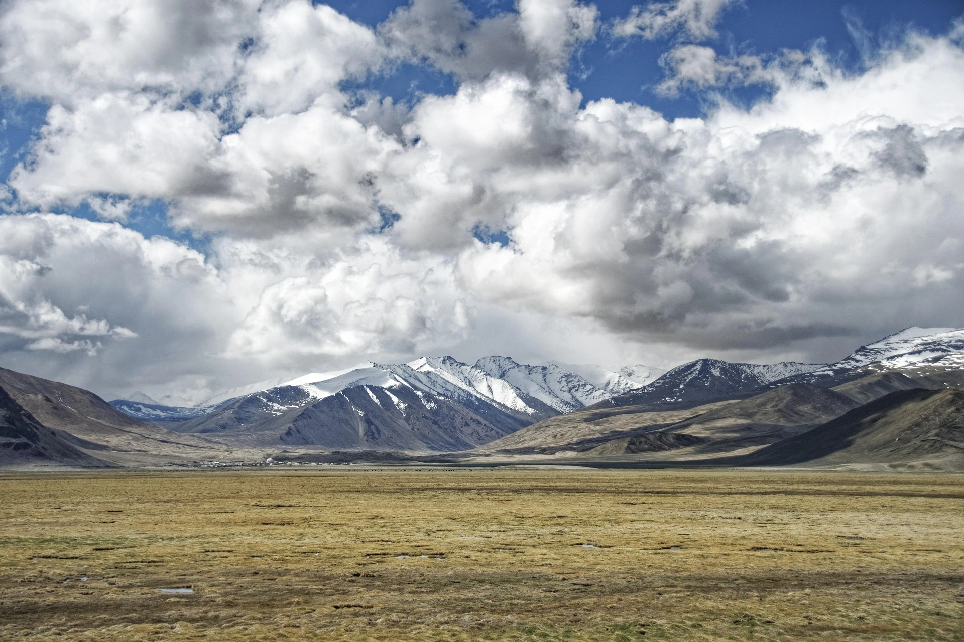 Wide alpine plateau with dry golden grass and snow-capped mountains beneath dramatic clouds.