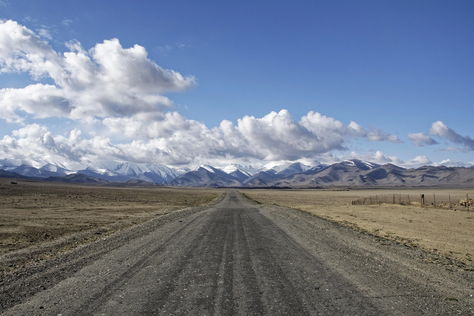 Lonely gravel road leading toward snow-capped Pamir mountains under a partly cloudy sky.