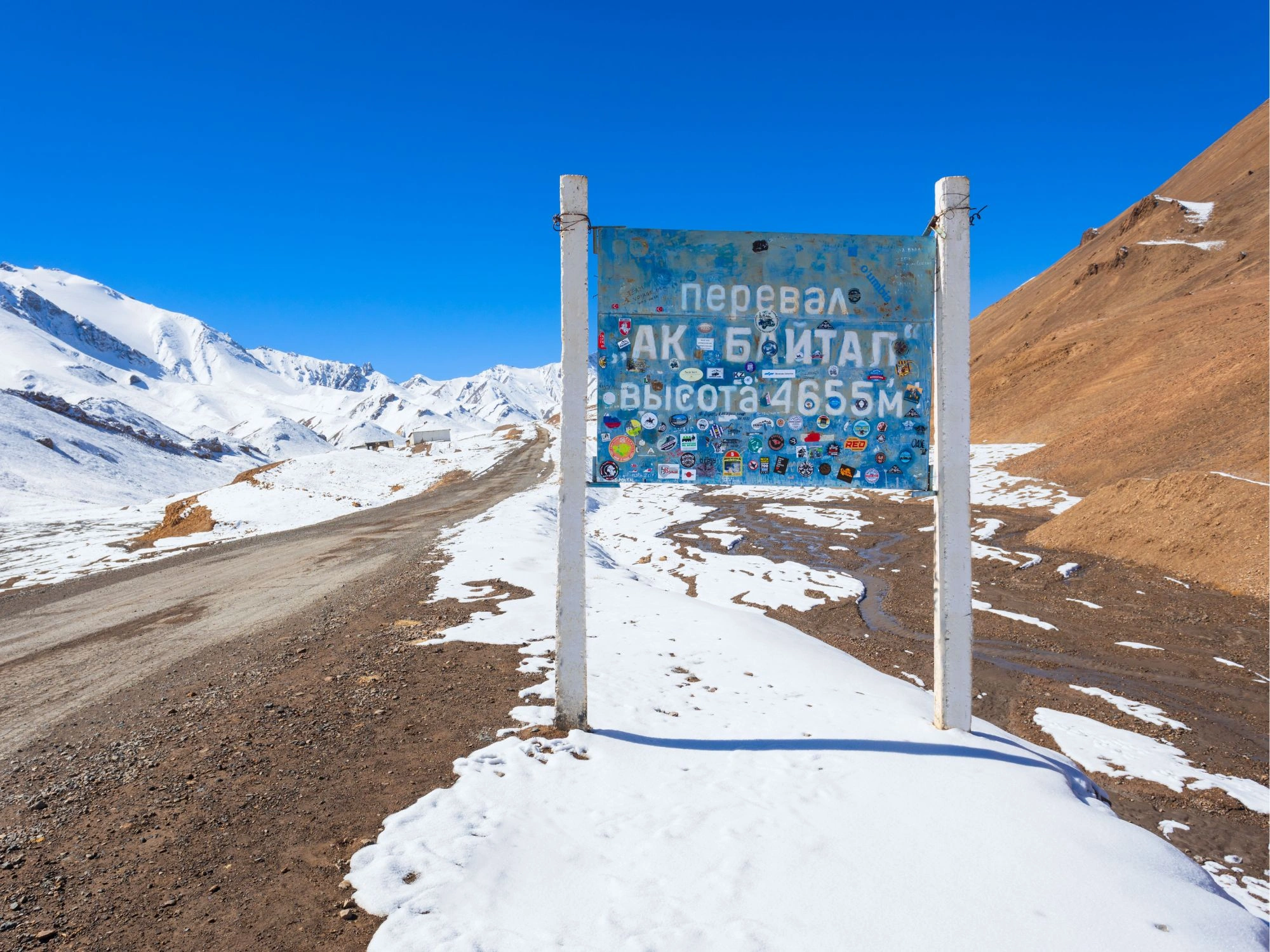 Ak-Baital Pass sign covered in stickers, marking 4,655m altitude on Tajikistan's Pamir Highway.