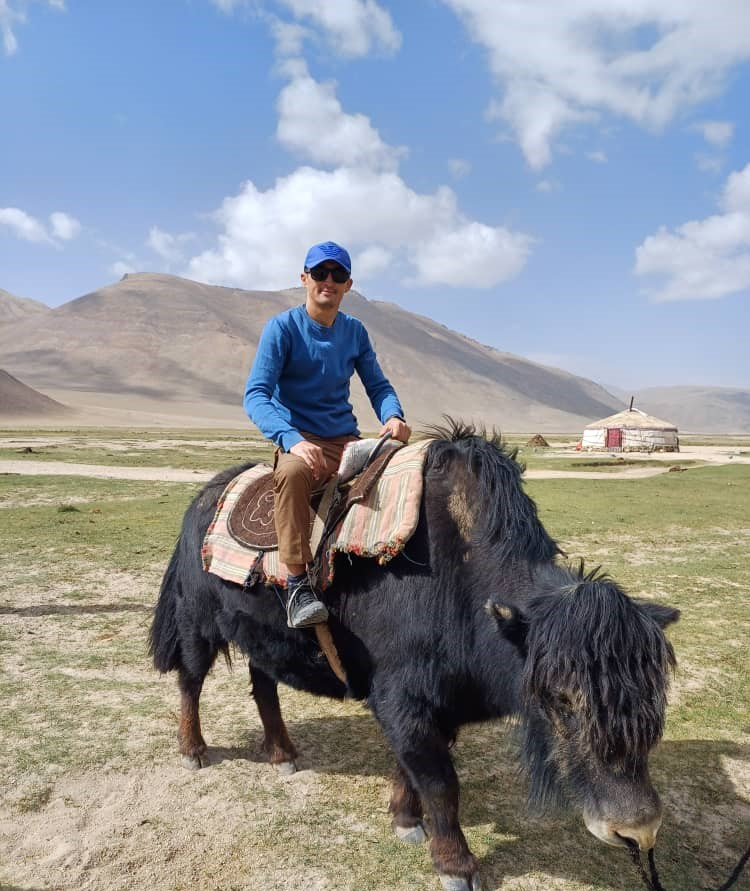 Traveler rides a yak on the Pamir Plateau in Tajikistan, with yurts and mountains in the background.