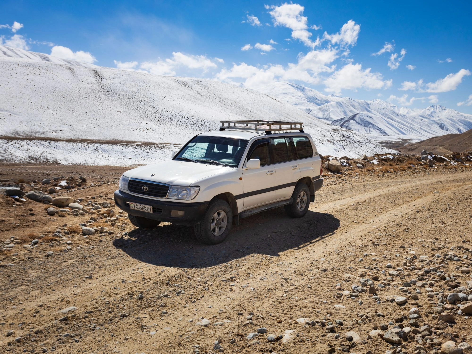 Toyota Land Cruiser on snowy mountain road near the Pamir Highway, under bright blue Tajik skies.