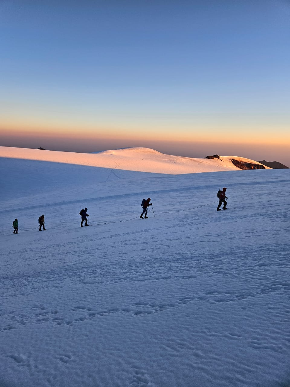 Rope team hikes across Mount Kazbek glacier under soft pastel sunrise sky.