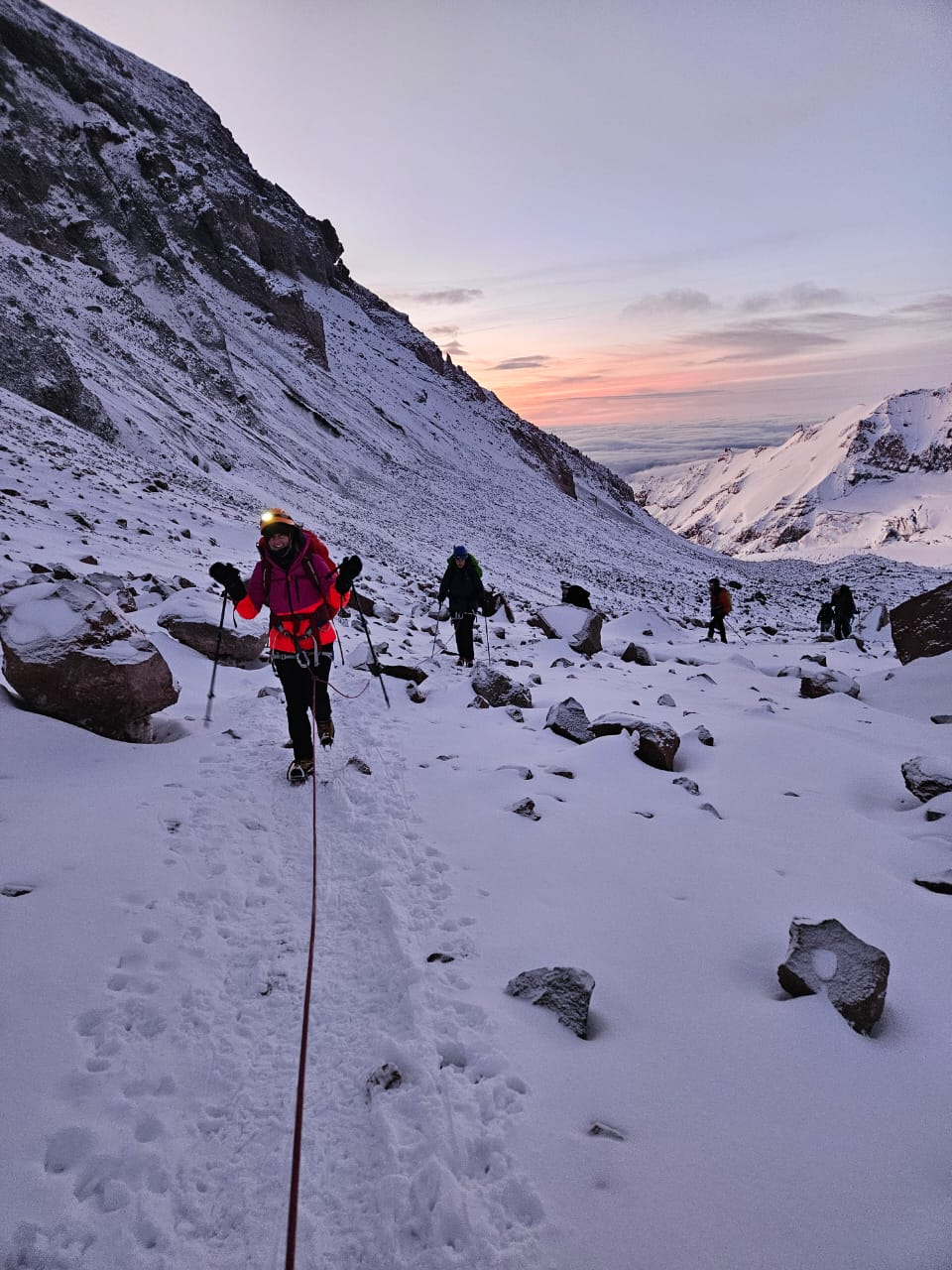 Climbers ascending snowy Mount Kazbek ridge at dawn with trekking poles and rope.