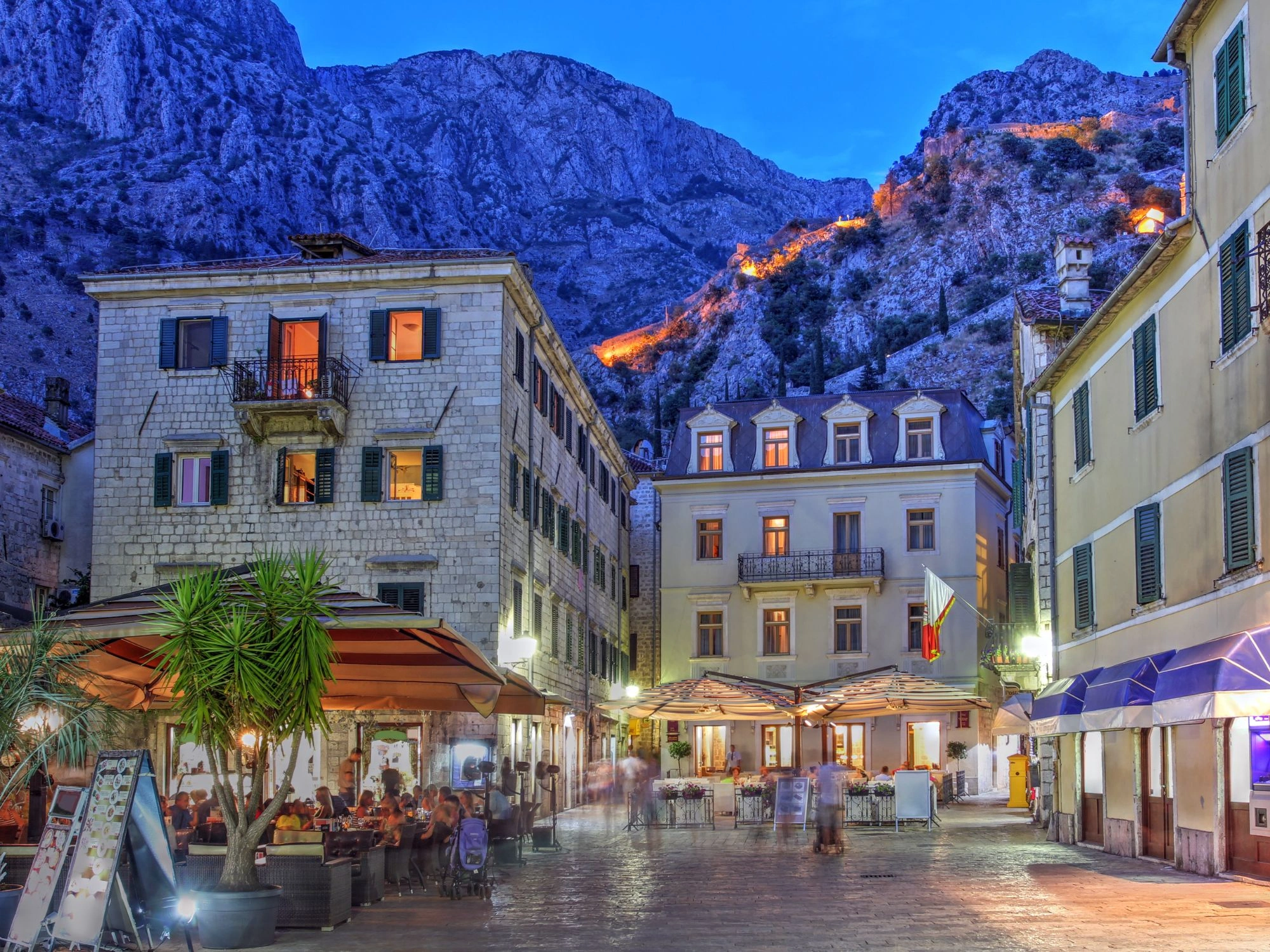 Kotor Old Town square at twilight with cozy cafés, historic stone buildings, and mountain backdrop, Montenegro.