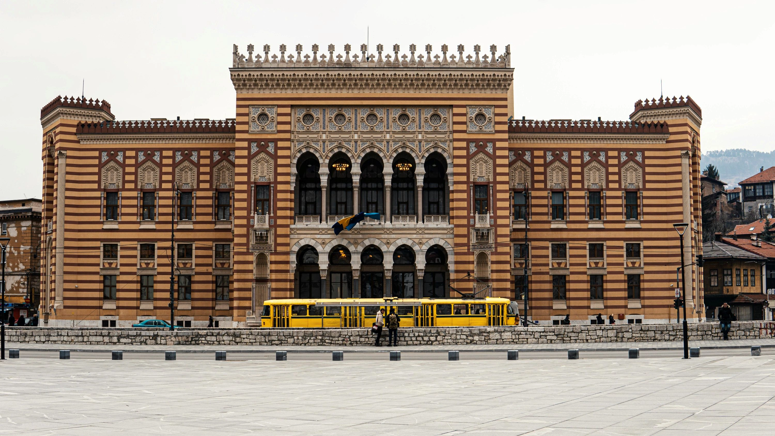 Sarajevo City Hall (Vijećnica) with Bosnian flag and a bright yellow tram in front.