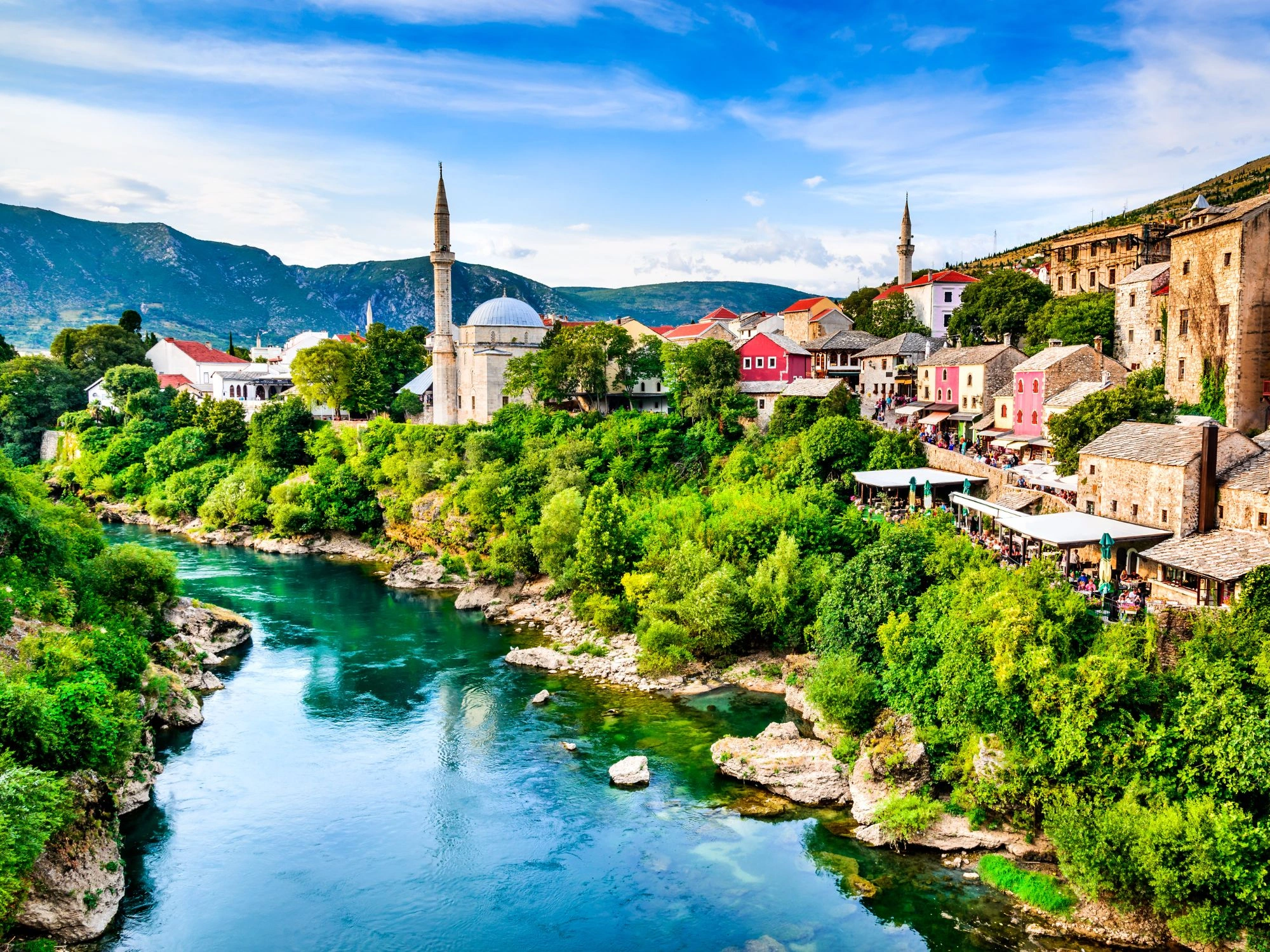 Colorful Old Town of Mostar, Bosnia, with mosques, river, and lush greenery under blue skies.