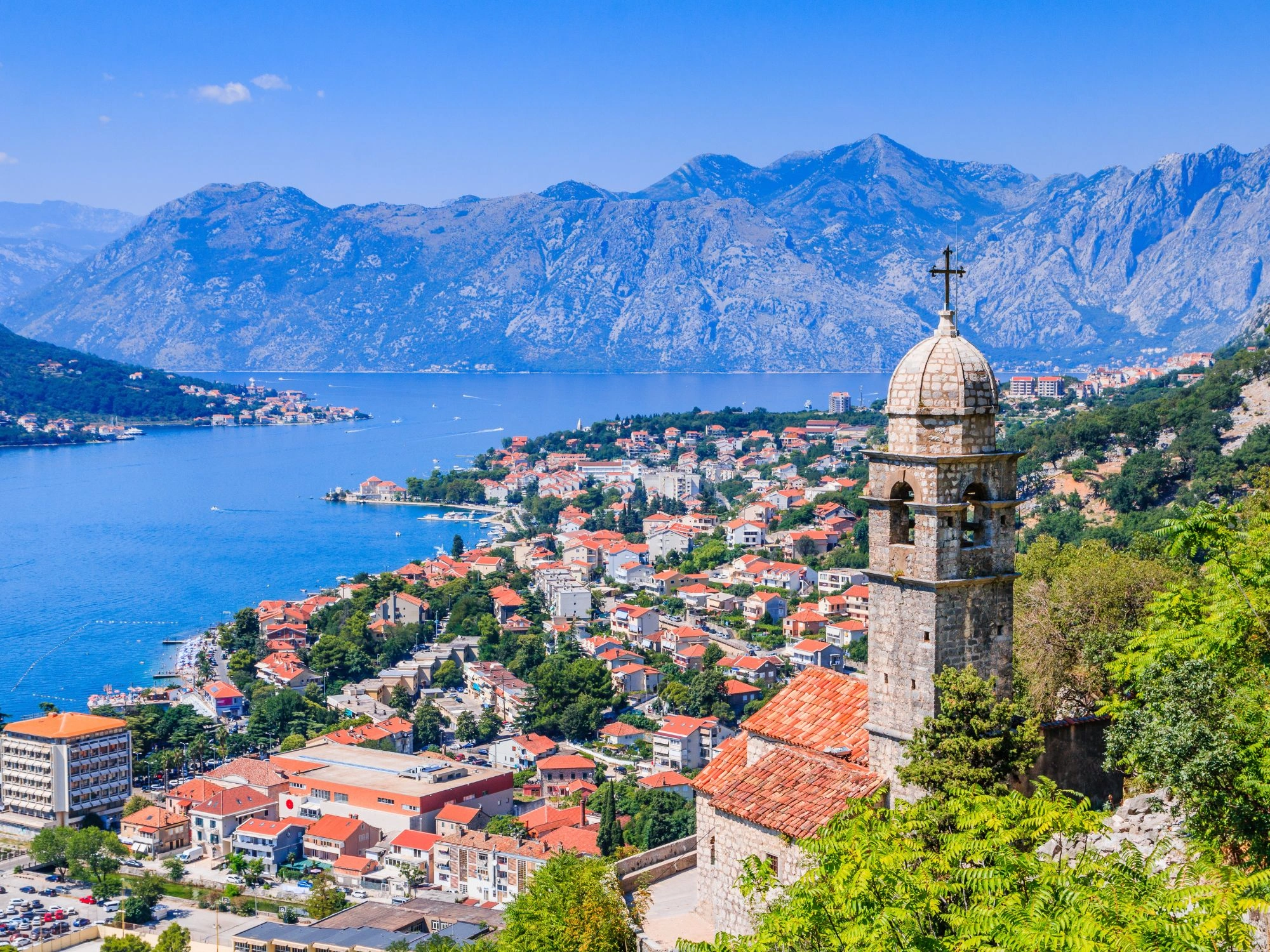 Scenic view of Kotor Bay and Old Town rooftops with a stone church tower, surrounded by mountains, Montenegro.