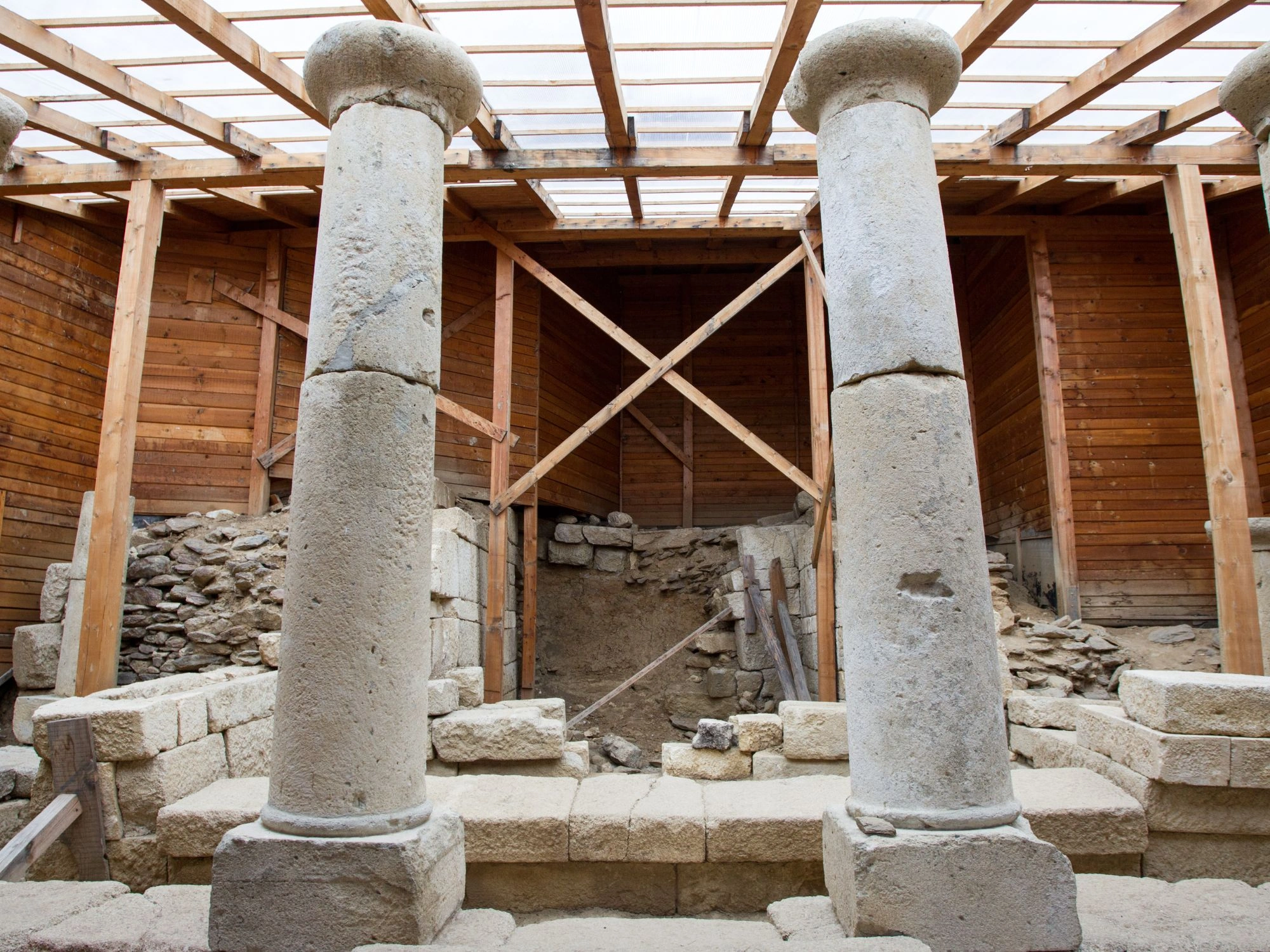 Ancient stone columns and ruins under wooden roof at an archaeological site.