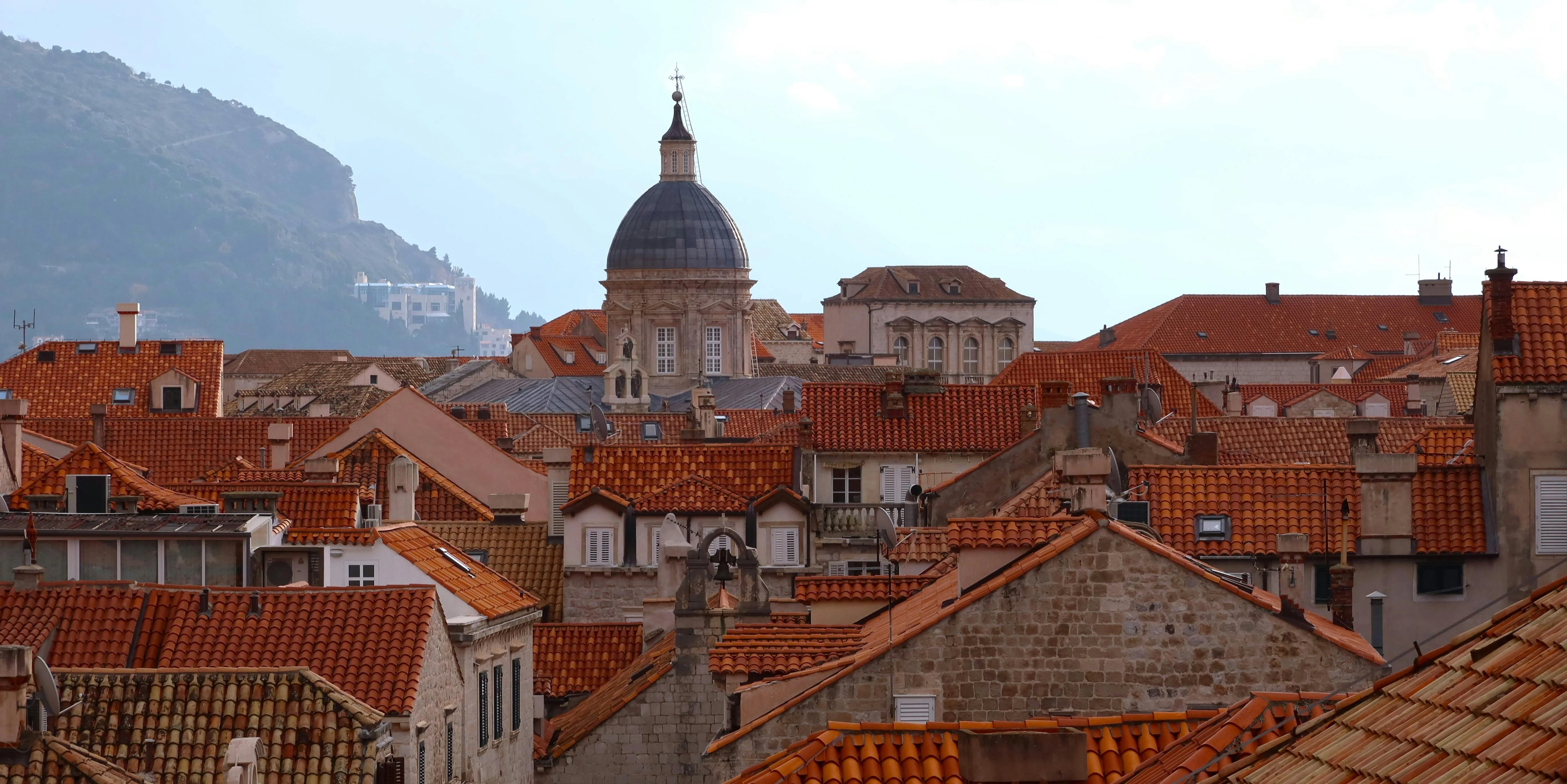 Orange-tiled rooftops of Dubrovnik Old Town with a cathedral dome and mountains in the distance.