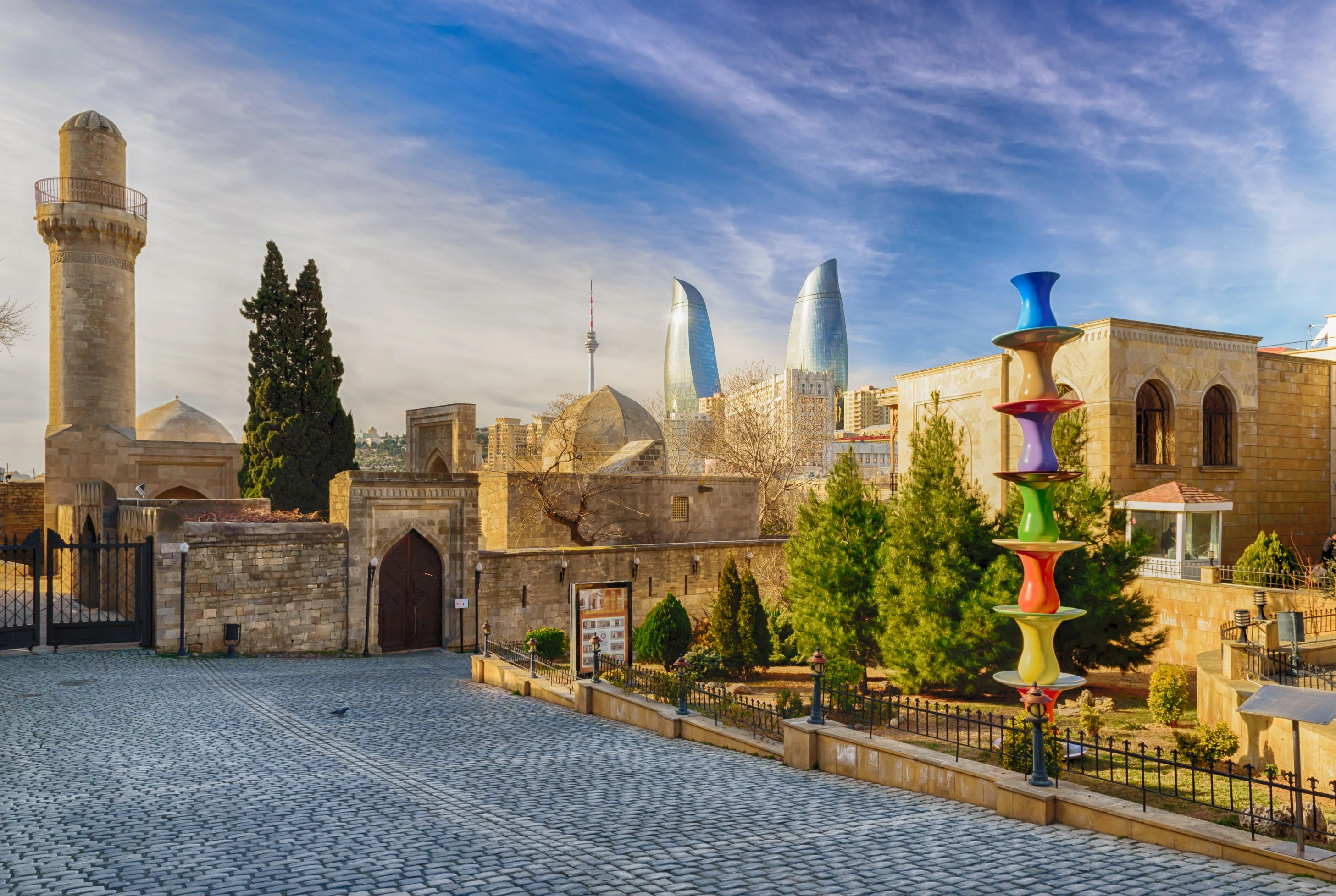 Shirvanshah Palace courtyard with Flame Towers in the background and a rainbow sculpture in front.