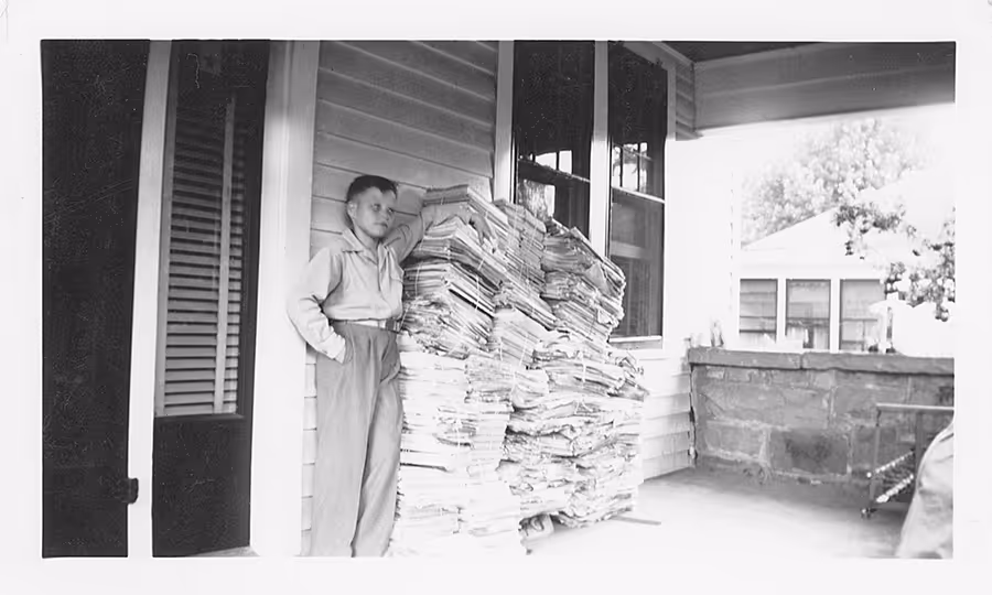 William J. O'Neil standing on his porch next to a stack of newspapers ready for him to deliver