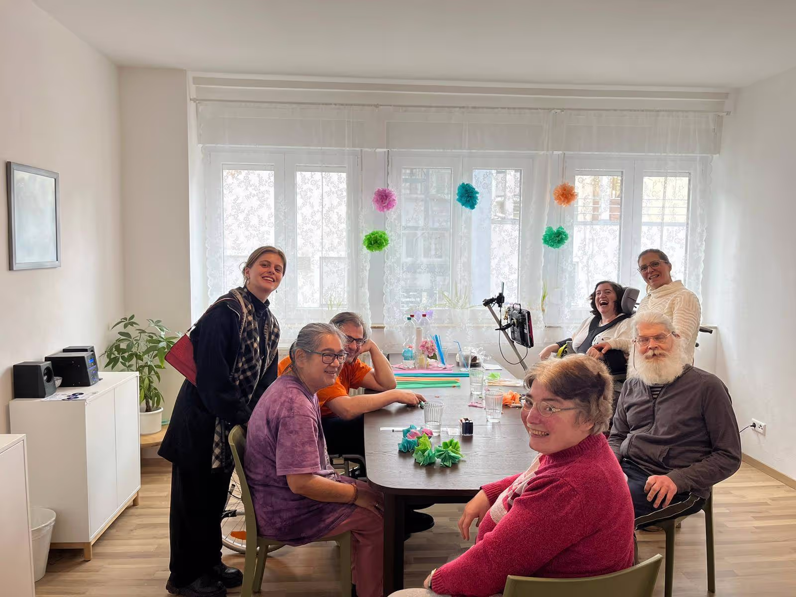 Mehrere Menschen sitzen gemeinsam in einem hellen Raum mit weißen Vorhängen und Blumen an einem Tisch und lächeln.