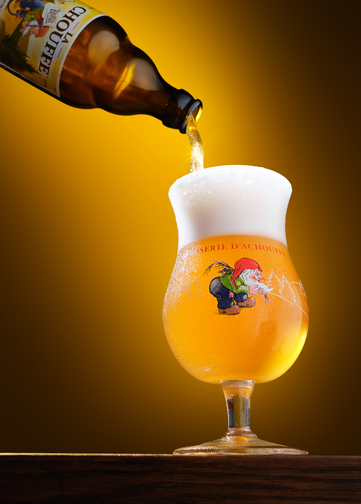 Golden La Chouffe beer being poured into a branded tulip glass with a thick white foam head.