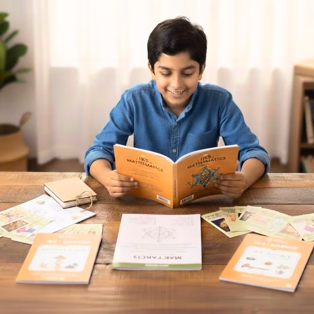 Smiling boy in blue shirt reading an IKS Mathematics book at a wooden table with other educational books and study cards.