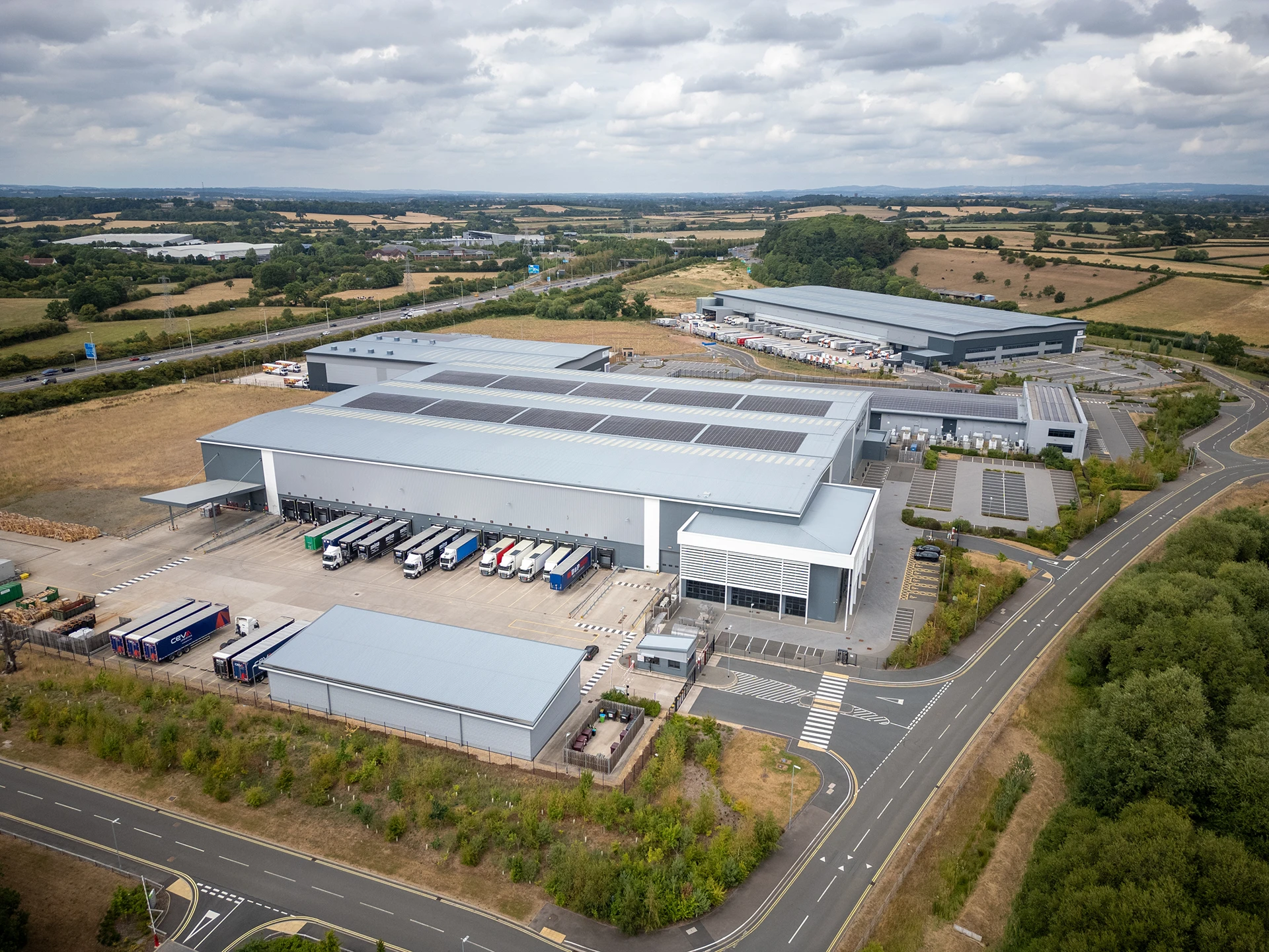 Large distribution warehouse with solar panels, multiple loading bays and trucks from aerial perspective