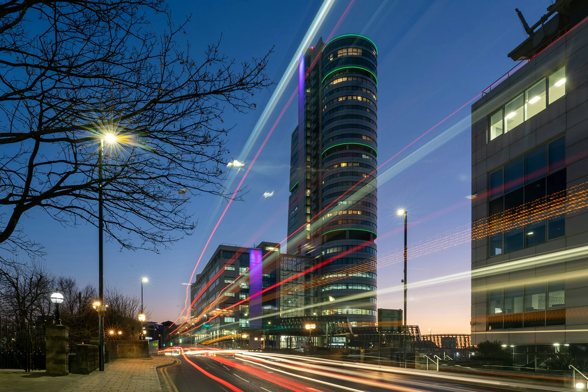 Tall skyscraper at night with colourful light trails from passing traffic.