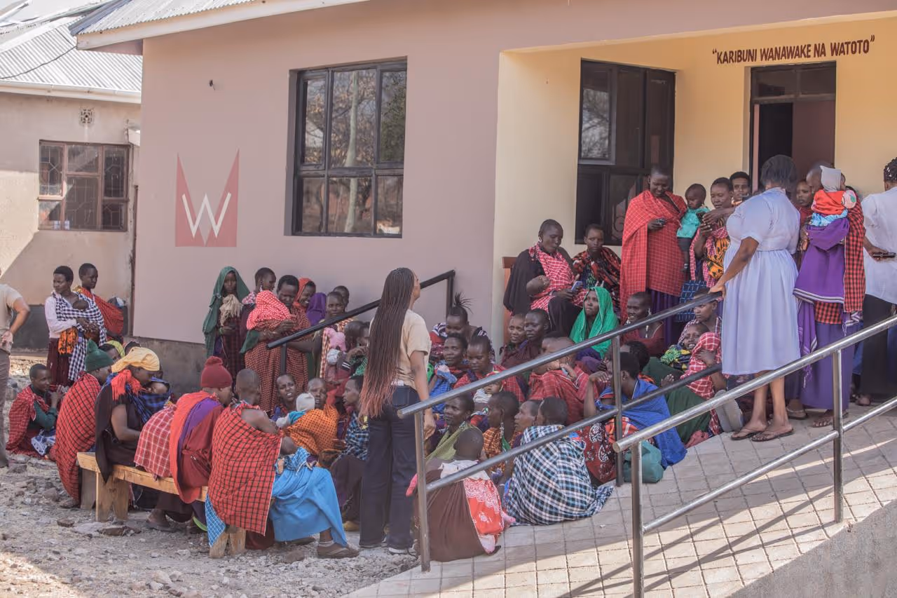 Group of women and children, many wearing colorful traditional Maasai shukas, gathered outside a building with a sign that reads 'KARIBUNI WANAWAKE NA WATOTO'.