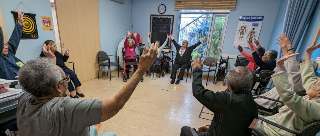 Group of elderly people seated in a circle in a room, raising their arms as part of a group exercise session.