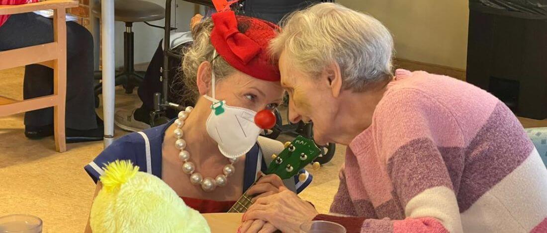 Person wearing a red hat, a mask with a red clown nose, and pearl necklace playing a green ukulele, engaging closely with an elderly person in a pink and purple sweater.