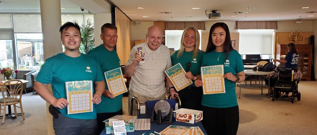 Group of four smiling volunteers wearing teal CWB shirts holding bingo cards with an elderly man holding a chocolate bar and standing behind a table with bingo supplies.