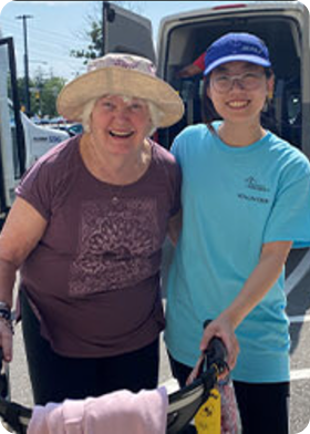 Smiling elderly woman wearing a hat and a younger woman in a blue shirt and cap standing together outdoors near an open vehicle door.