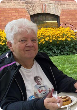 Elderly woman with white hair sitting outdoors holding a fork and a plate with a cinnamon roll, in front of yellow flowers and a brick building.